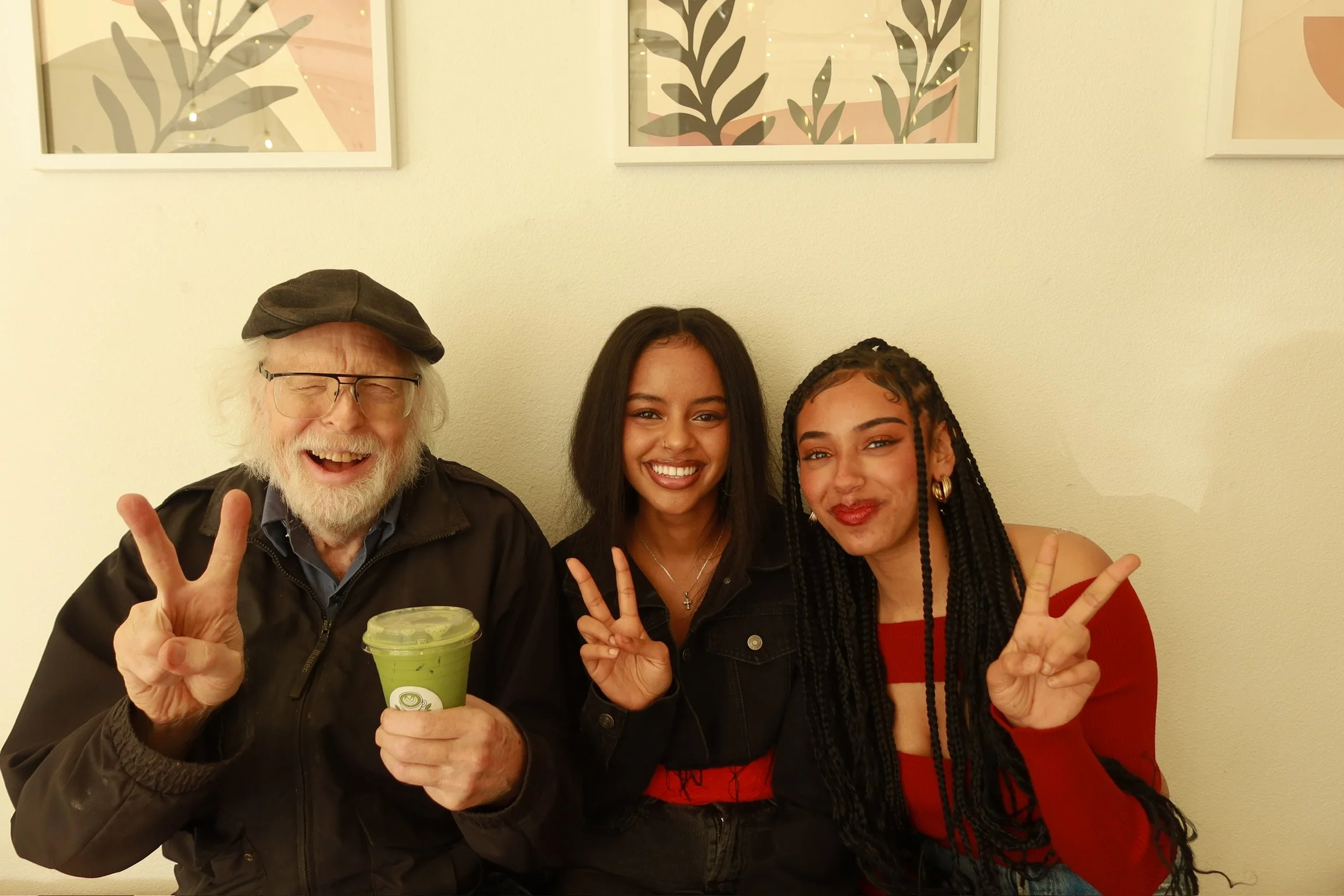 Three people smiling and making peace signs in front of a light-colored wall with framed art. The man on the left has a beard, glasses, and a brown cap, holding a green beverage. The two women on the right have dark hair, with one wearing a black jac