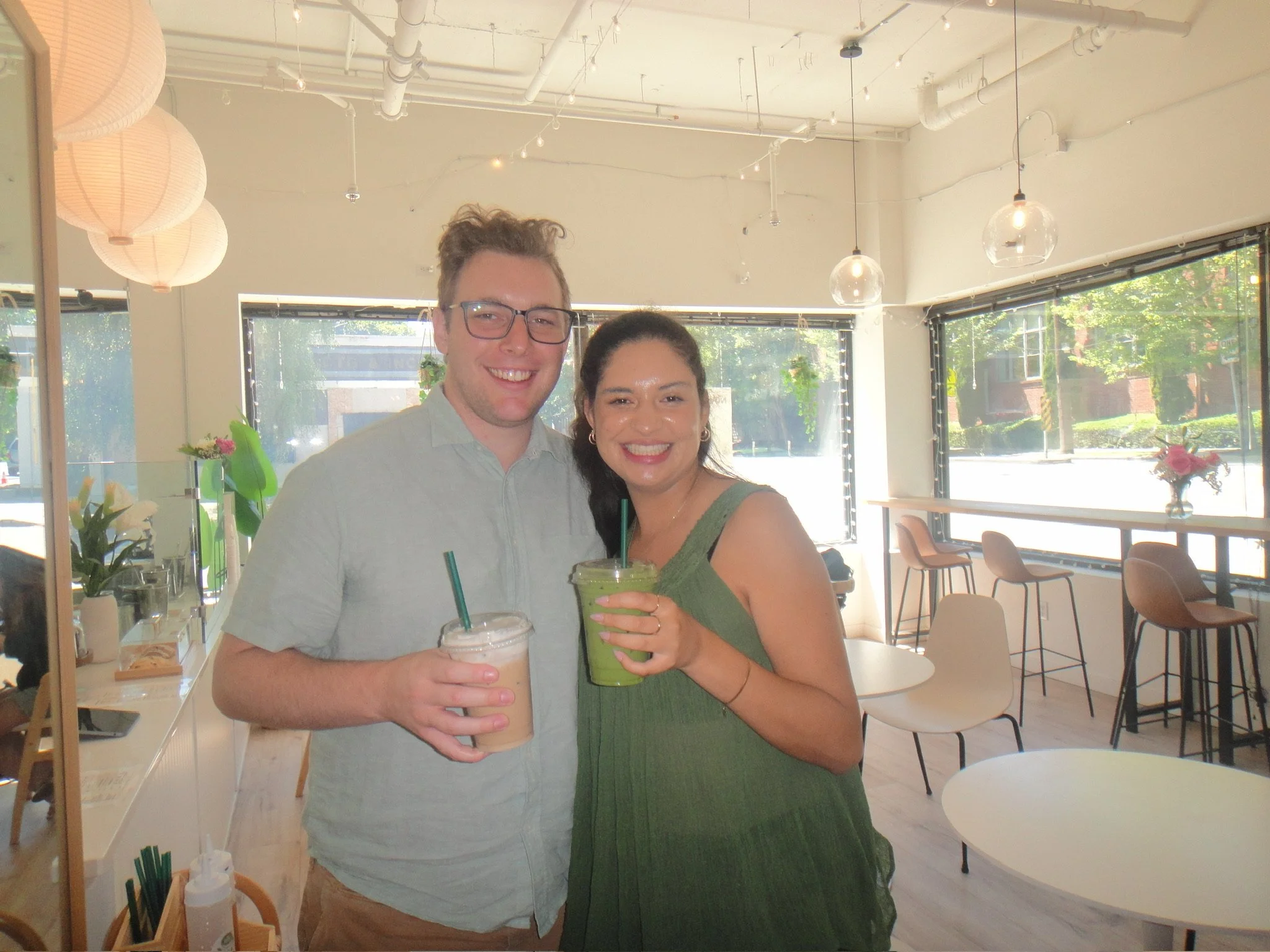 A smiling young man and woman holding drinks inside a modern cafe or restaurant with large windows, hanging lights, and minimalistic decor.