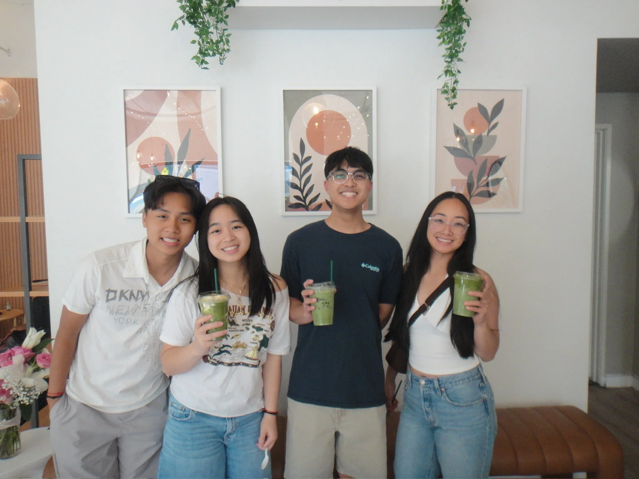 Four young adults standing together and smiling indoors, holding green drinks, with framed abstract art and green hanging plants on the wall behind them.
