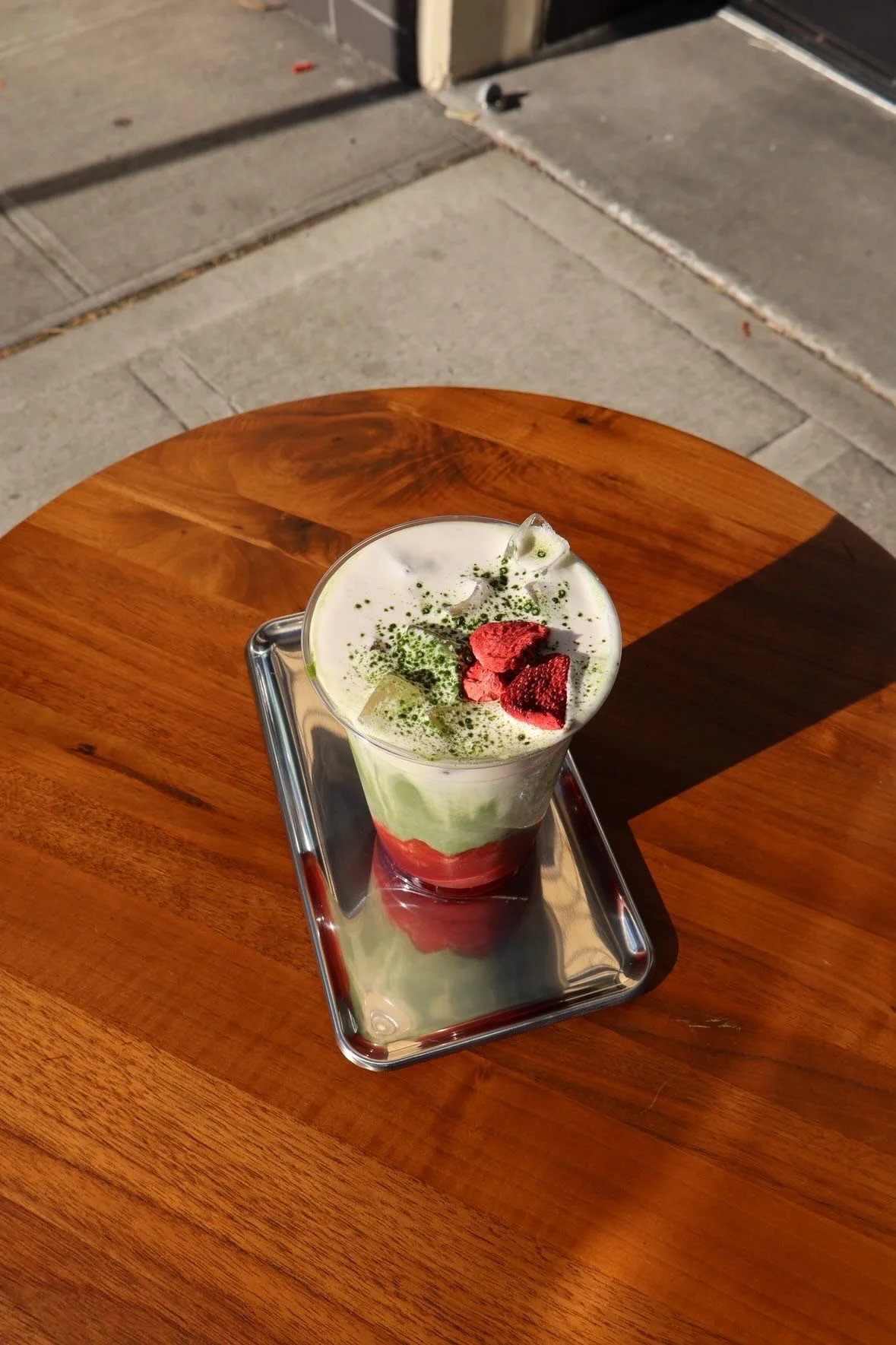 A layered green and red dessert topped with strawberries and green powder, served in a tall glass on a metal tray on a wooden table, with an outdoor background.