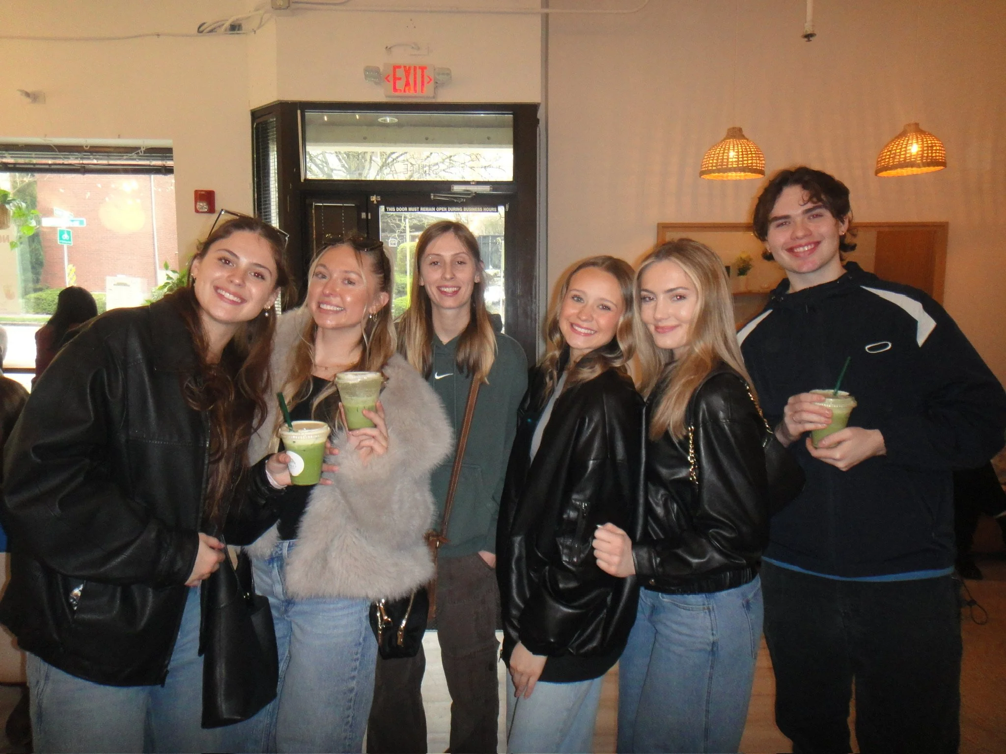 Six young adults smiling and posing for a photo inside a café, holding green smoothies.