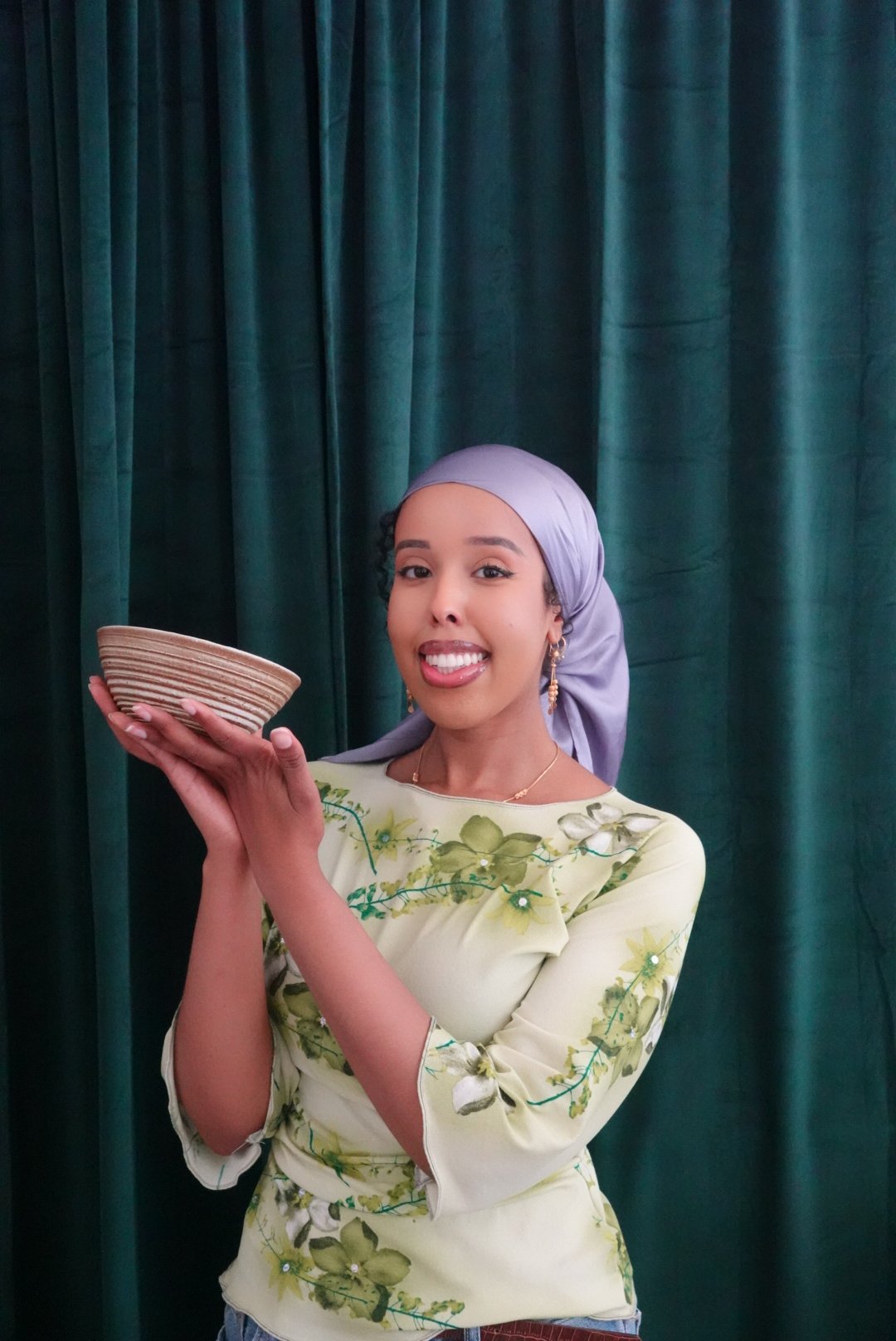 A young woman wearing a lavender headscarf and floral yellow top holding a striped ceramic bowl in front of a green curtain.