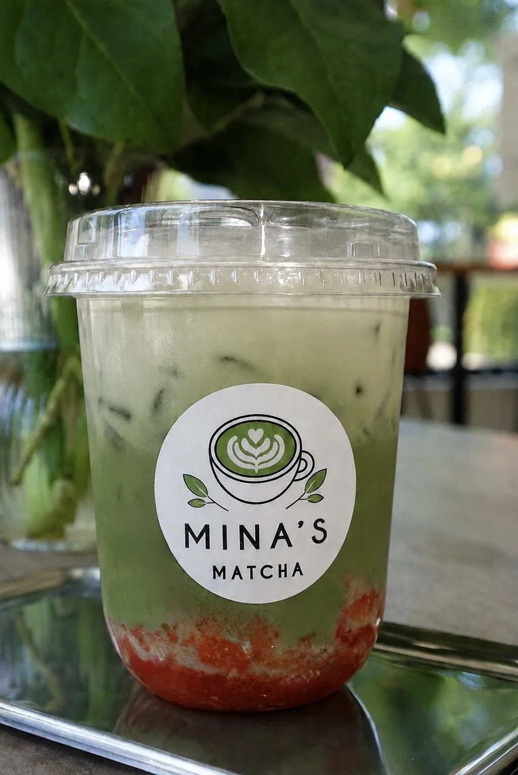 A plastic cup with a clear lid containing a layered matcha green tea drink topped with strawberry syrup, labeled 'Mina's Matcha' with a logo of a latte art heart in a green matcha cup surrounded by leaves, placed on a reflective surface outdoors.