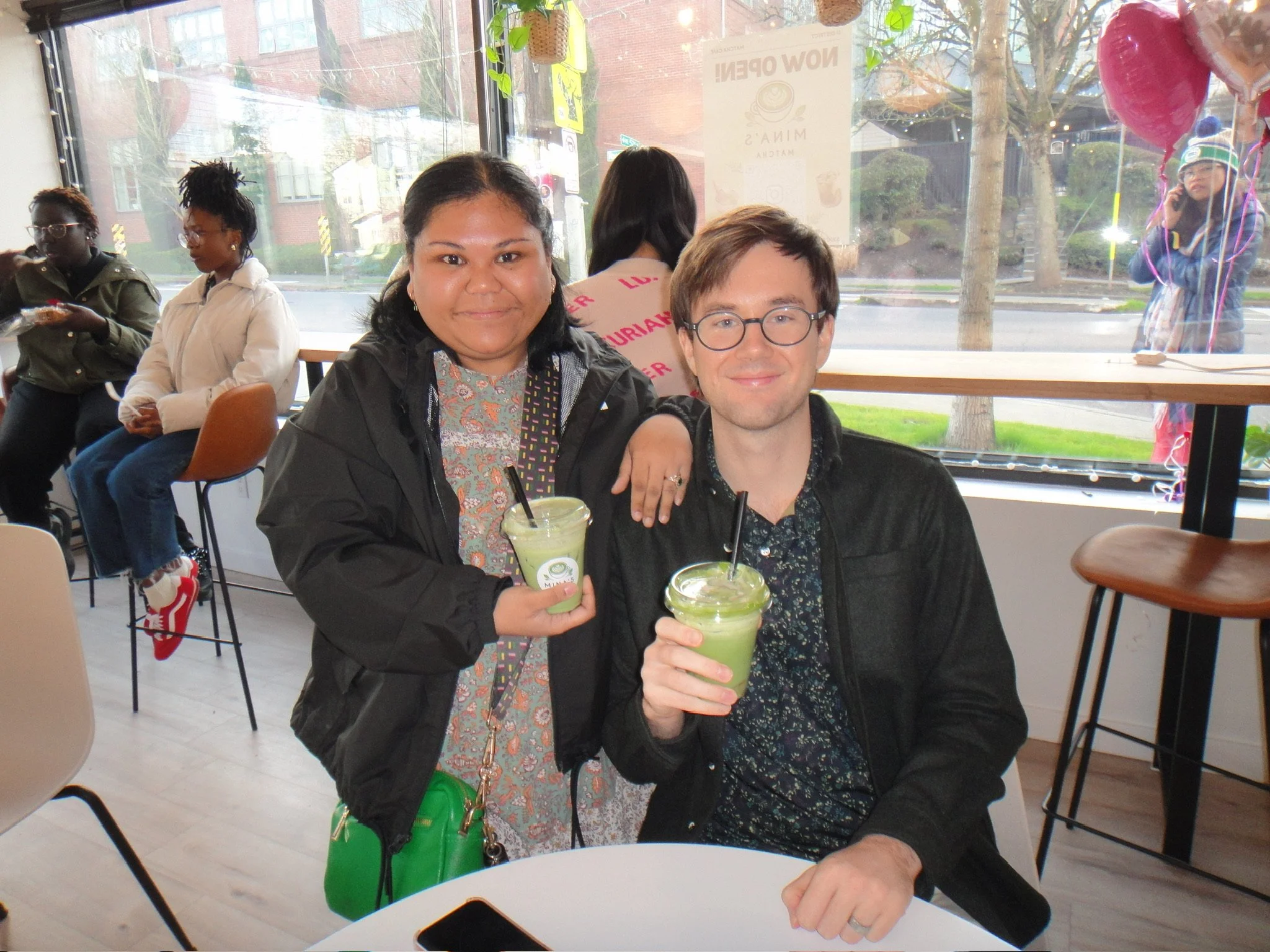 Two people sitting at a cafe table, holding green smoothies and smiling at the camera, with others in the background inside a cafe with large windows.