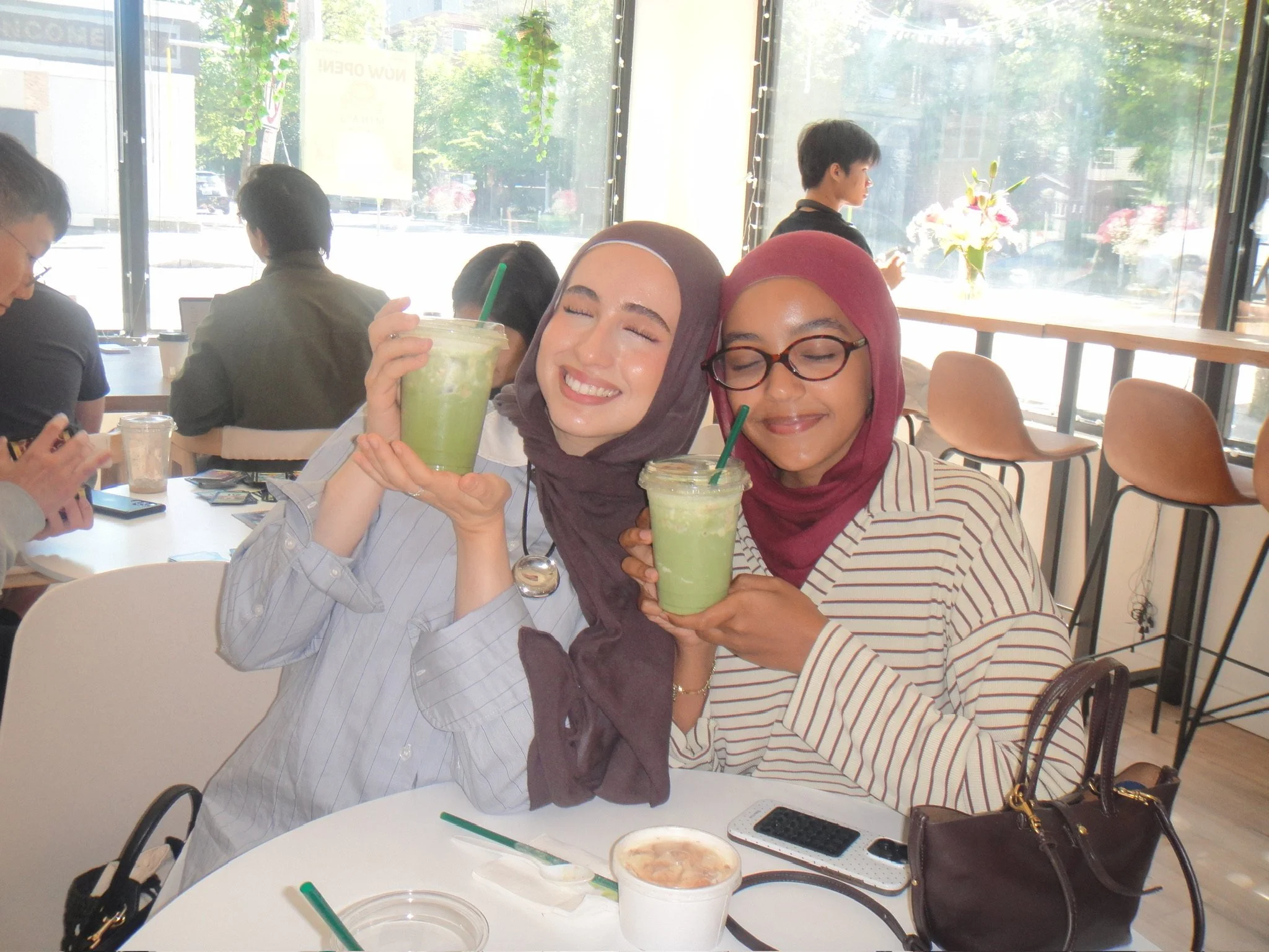Two women wearing hijabs sitting at a table in a cafe, holding green smoothies and smiling with their eyes closed, with other people and large windows in the background.