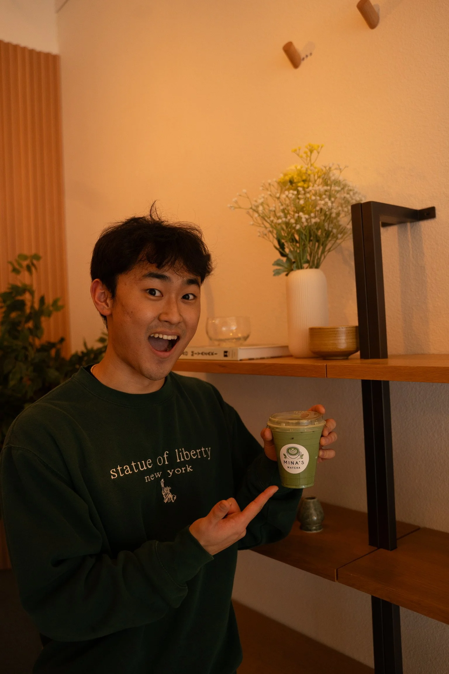 A young man with dark hair smiling and holding a green matcha drink in a clear cup with a label that reads 'Mina's Matcha.' He is wearing a dark sweatshirt with text that reads 'statue of liberty new york.' The background features a wooden shelf with