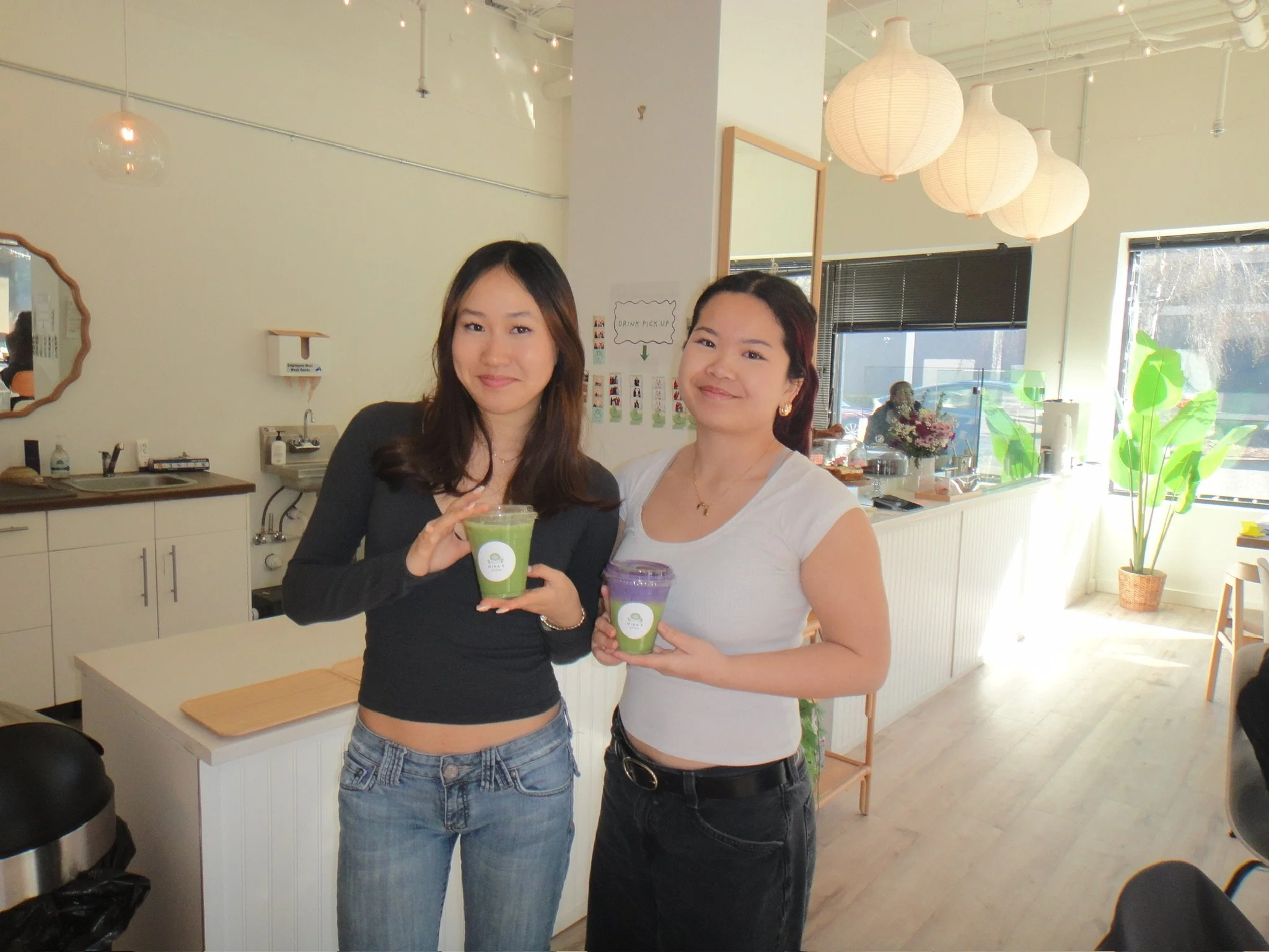 Two women standing inside a bright, modern cafe, smiling and holding colorful drinks. The woman on the left has long brown hair, wearing a black top and jeans. The woman on the right has dark hair with a reddish tint, wearing a white top, and earring