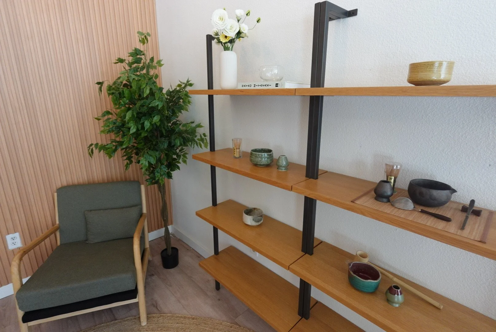 A corner of a room with a green armchair beside a tall potted plant. Two wooden and black metal shelves display various ceramic and glass decorative items, including vases, bowls, and cups. The wall behind the shelves is white, with a section of vertical wooden paneling on the left side.