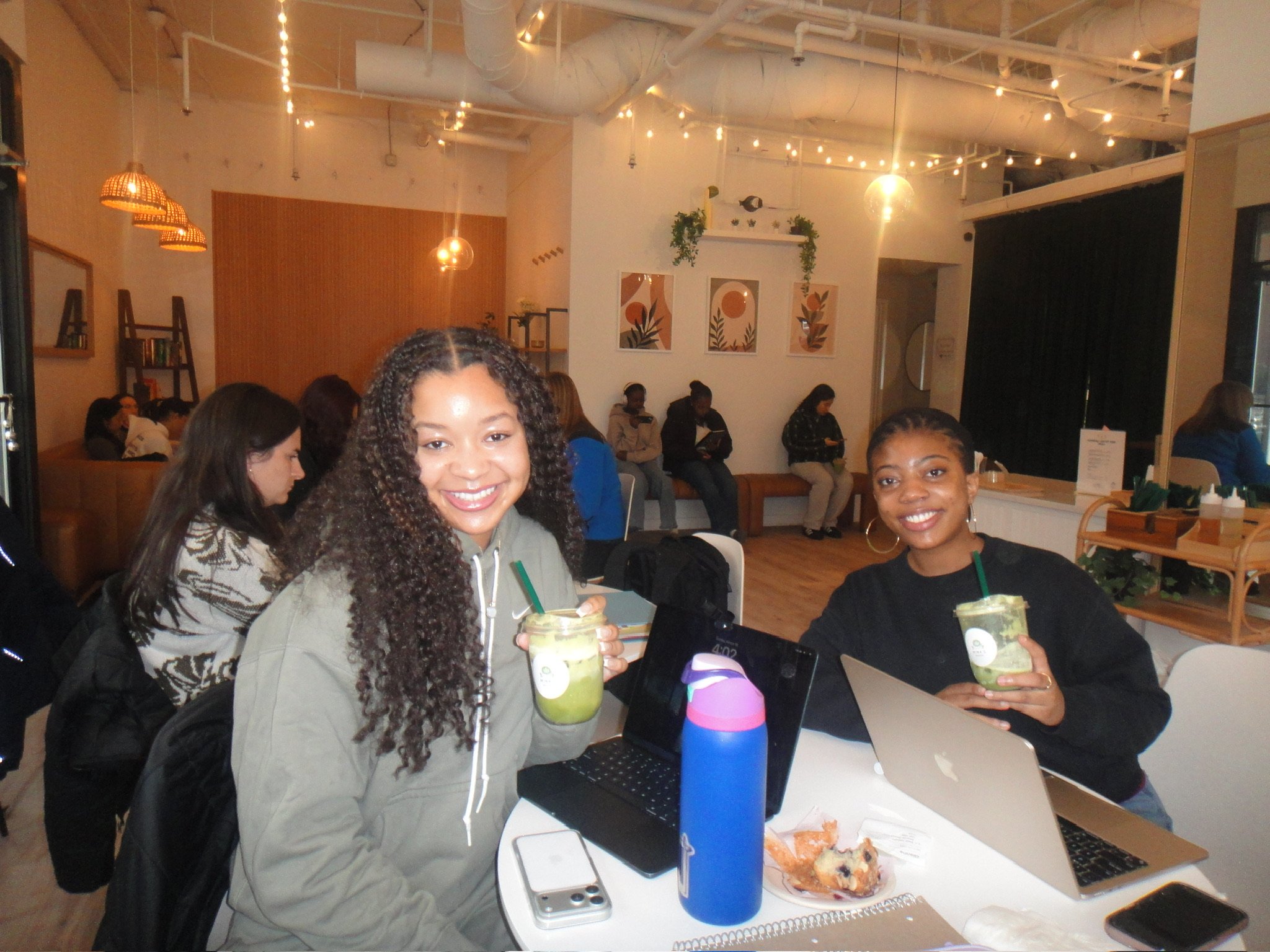 Two women sitting at a table in a cafe, smiling at the camera, holding green drinks, with laptops and a pizza on the table. Other people are seated in the background.