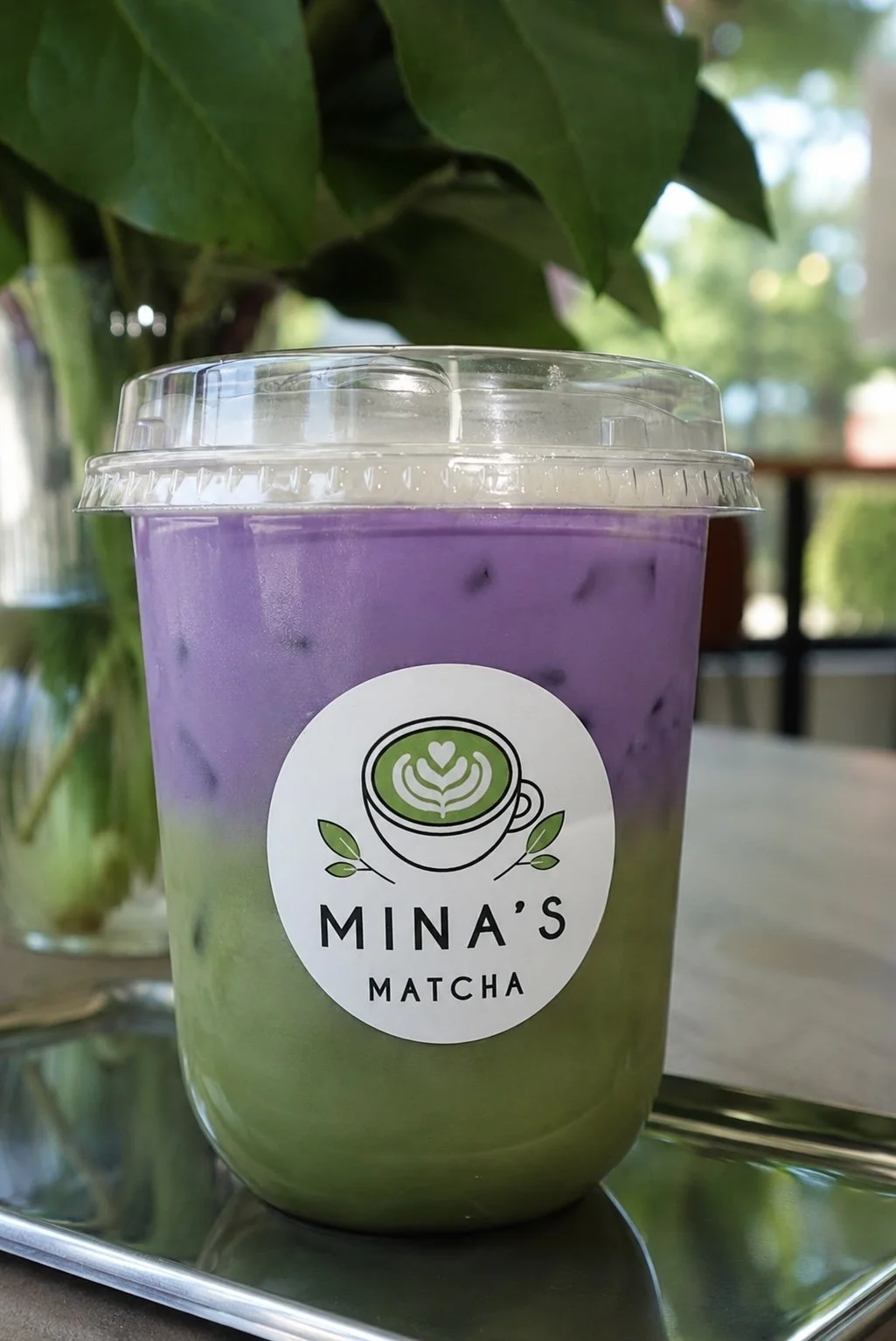 A plastic cup of purple ube layered matcha latte with ice, branded with Mina's Matcha logo, in front of green leaves.