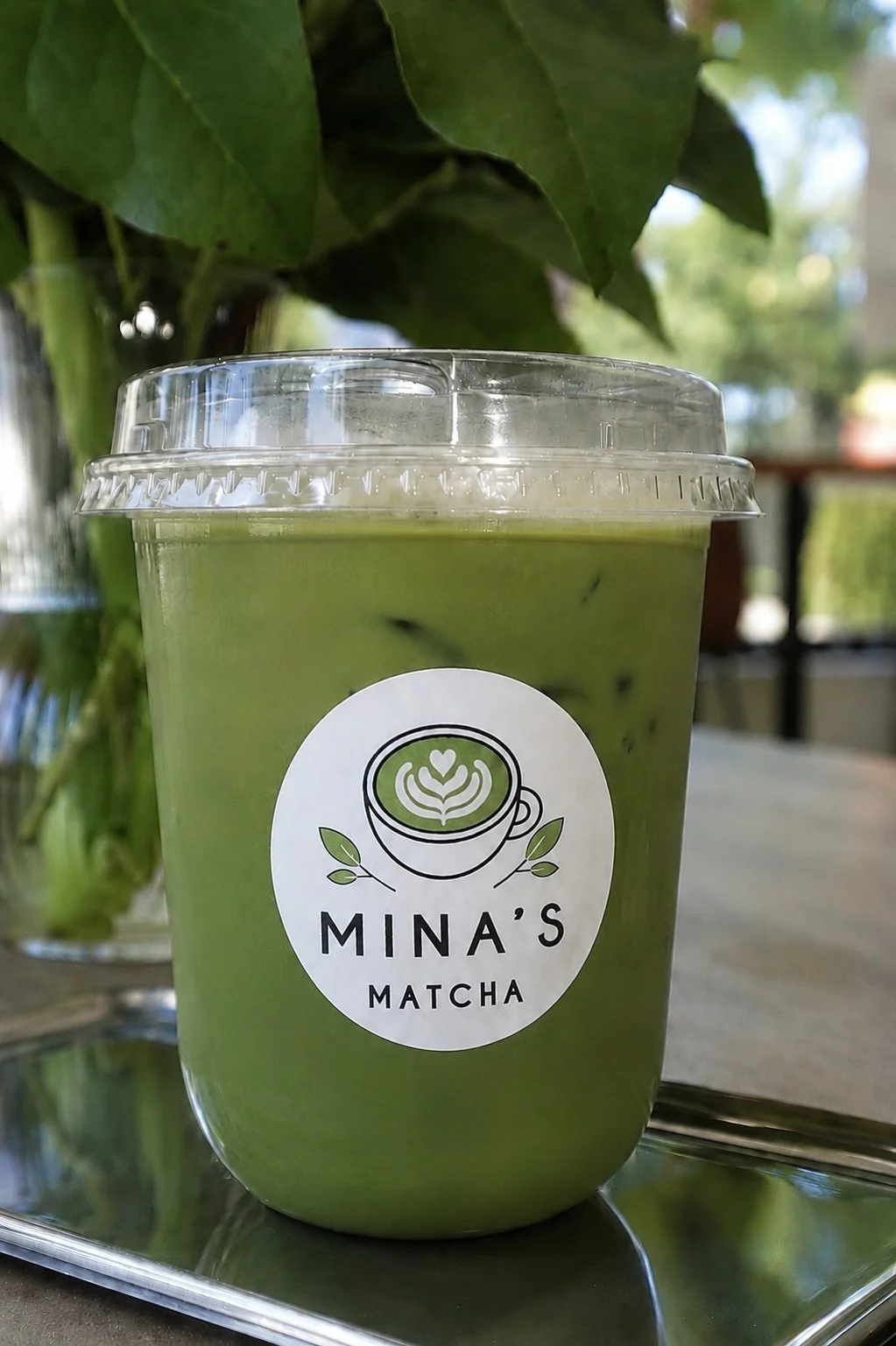 A plastic cup filled with green matcha latte, featuring a logo with a latte art heart and leaves, from Mina's Matcha, on a reflective tray.