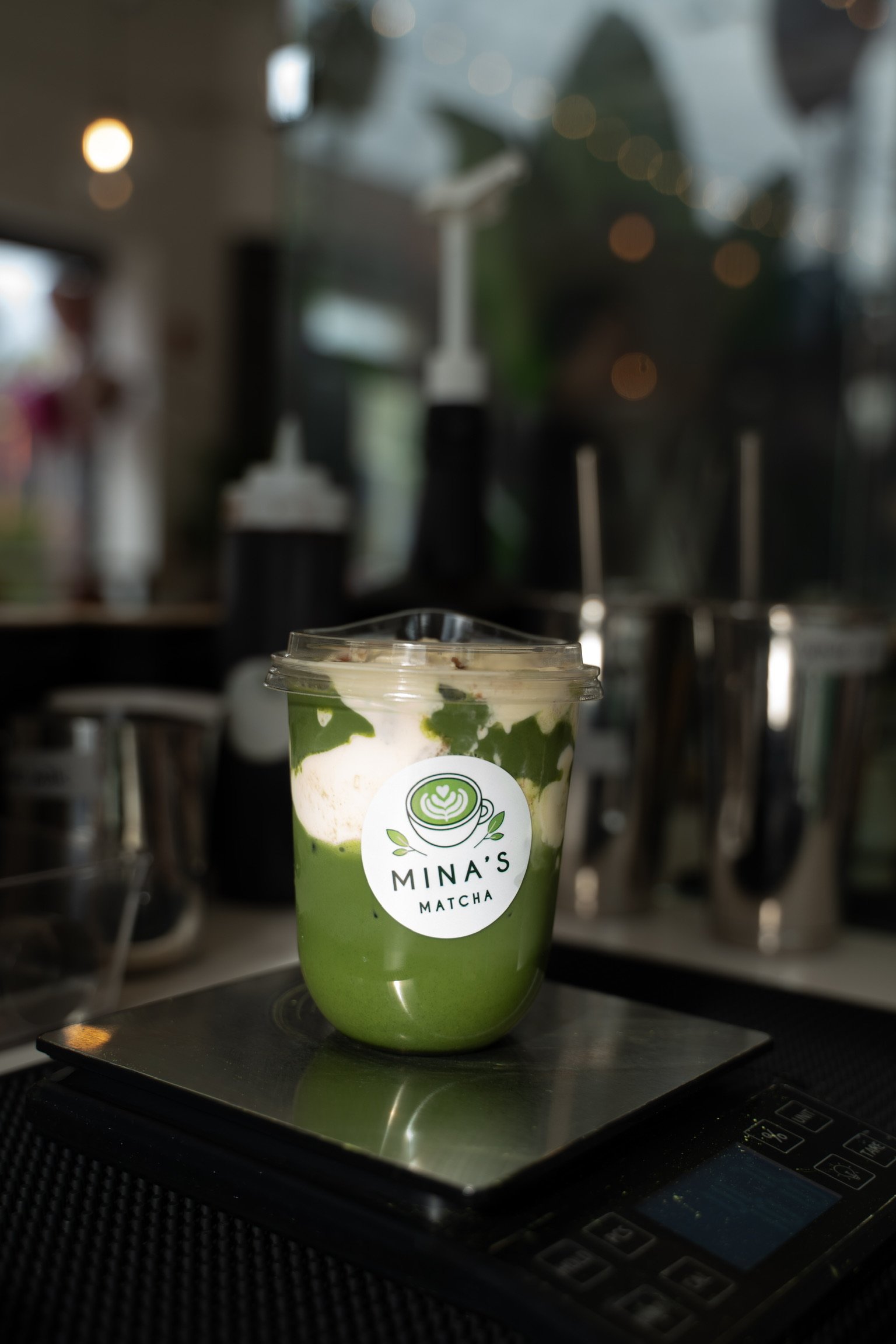 A matcha latte in a plastic cup with a clear lid on a scale, with the logo of Mina's Matcha on the cup, inside a coffee shop or cafe.