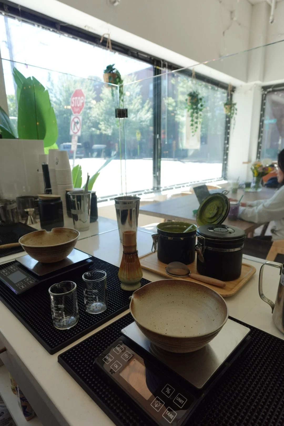 Indoor scene at a cafe or restaurant with tables, bowls, glasses, and food containers. Large window with outdoor view, plants, stop sign, and a person using a laptop.