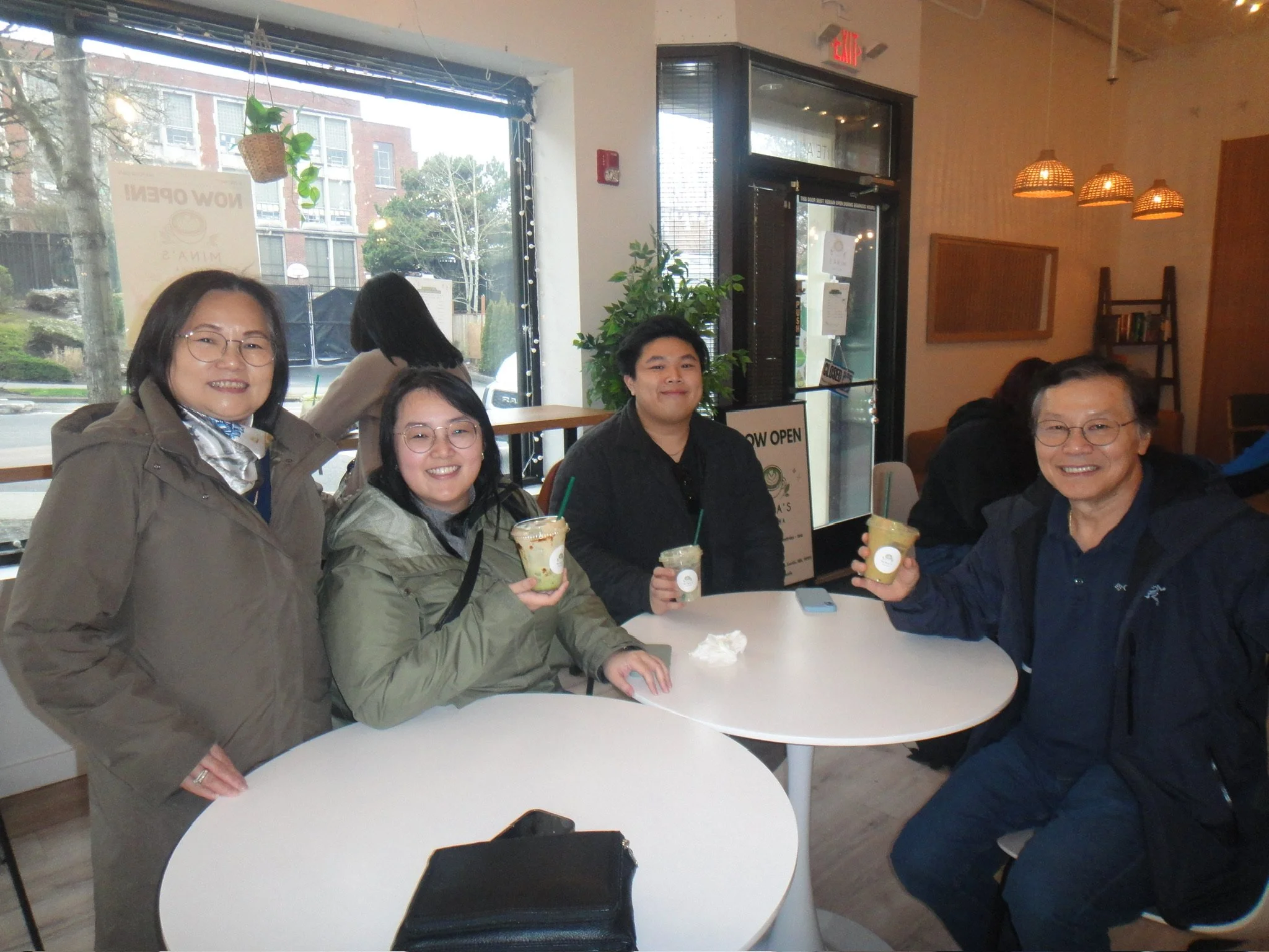 Four people sitting at a restaurant table, smiling and holding drinks, with a large window and indoor plants in the background.