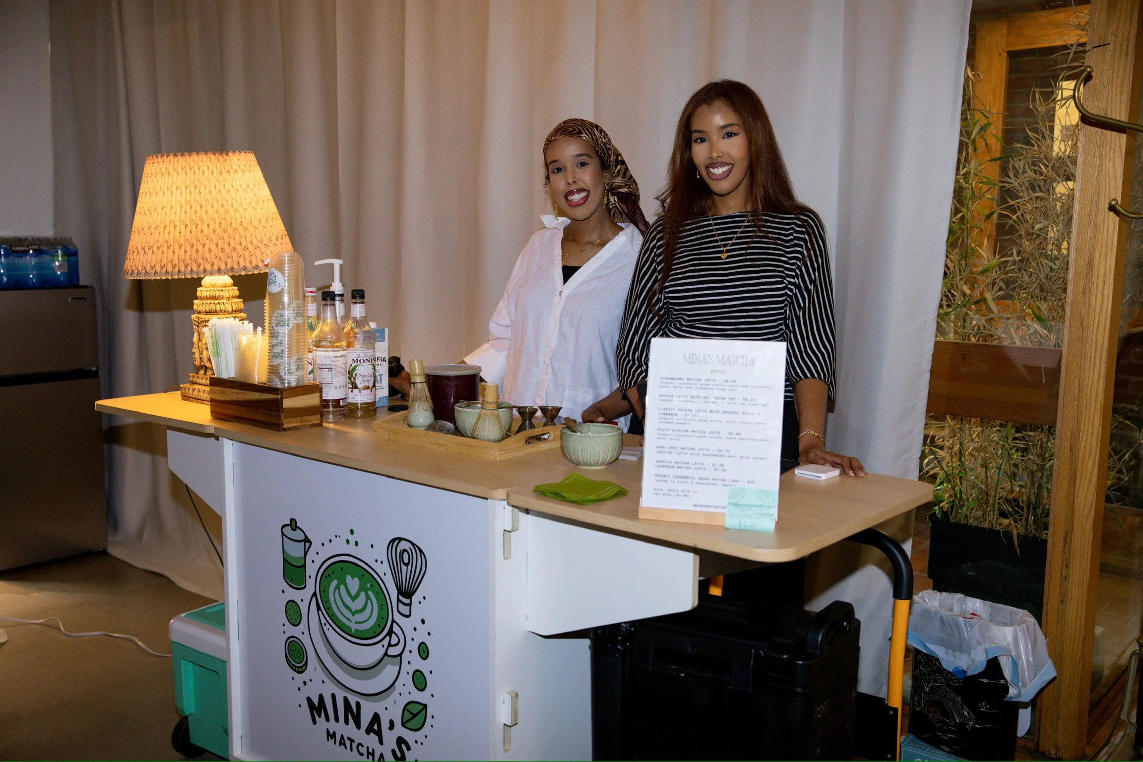 Two women stand behind a small stand with a white table at an indoor event. The stand has a colorful logo with leaves, cups, and the words "MINA'S MATCHA". One woman wears a striped shirt and has long, reddish-brown hair, smiling at the camera. The other woman wears a white shirt and a headscarf, also smiling. The table displays bottles, cups, a menu, and other items for serving matcha.