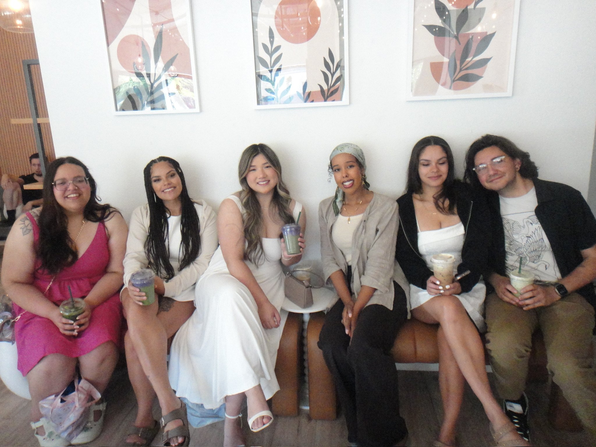 Group of six diverse women sitting together, smiling, holding drinks, indoors with framed artwork on the wall behind them.