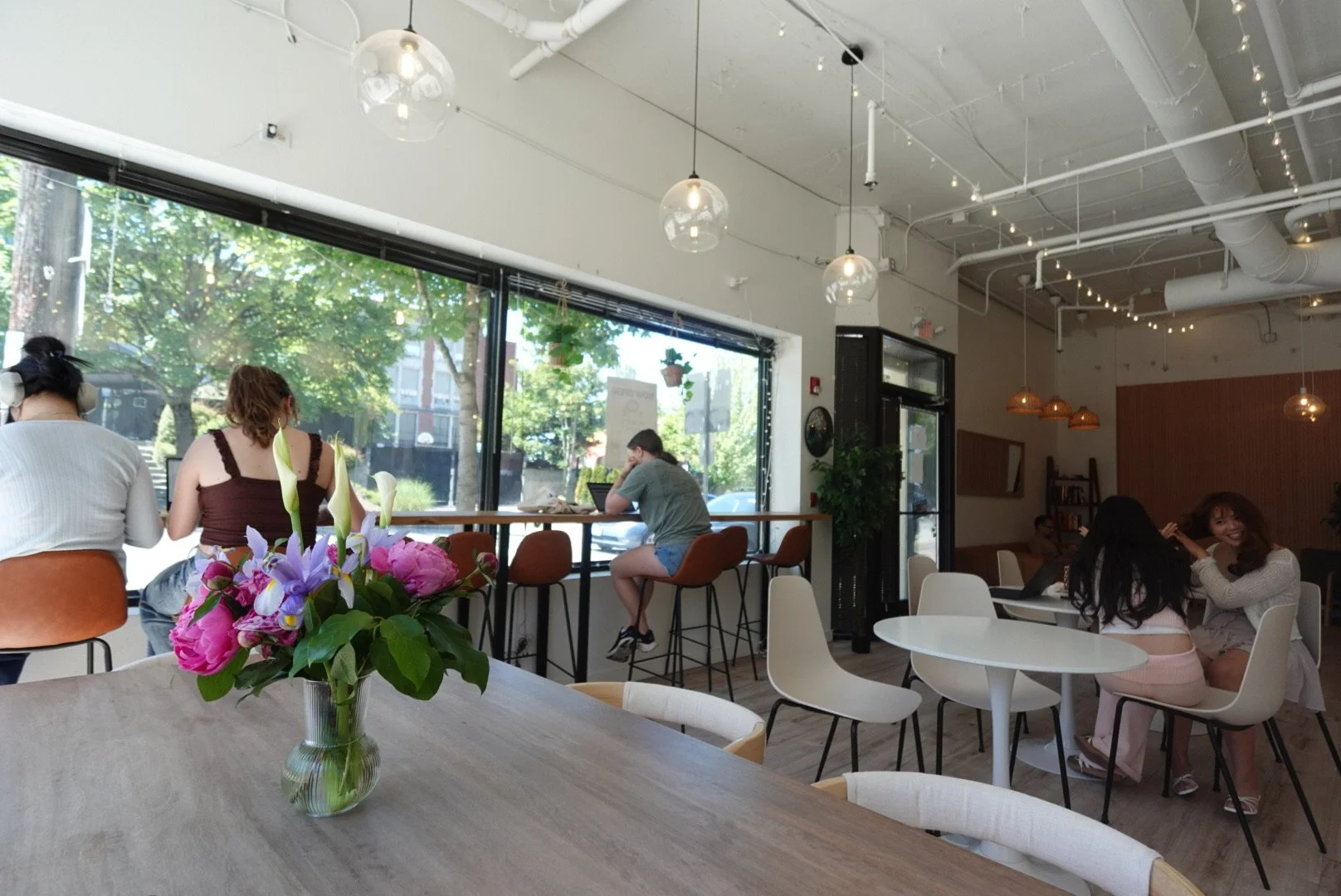 Interior of a bright, modern cafe with large windows, hanging pendant lights, and a small table with a vase of colorful flowers in the foreground. Several people are sitting and chatting or working.