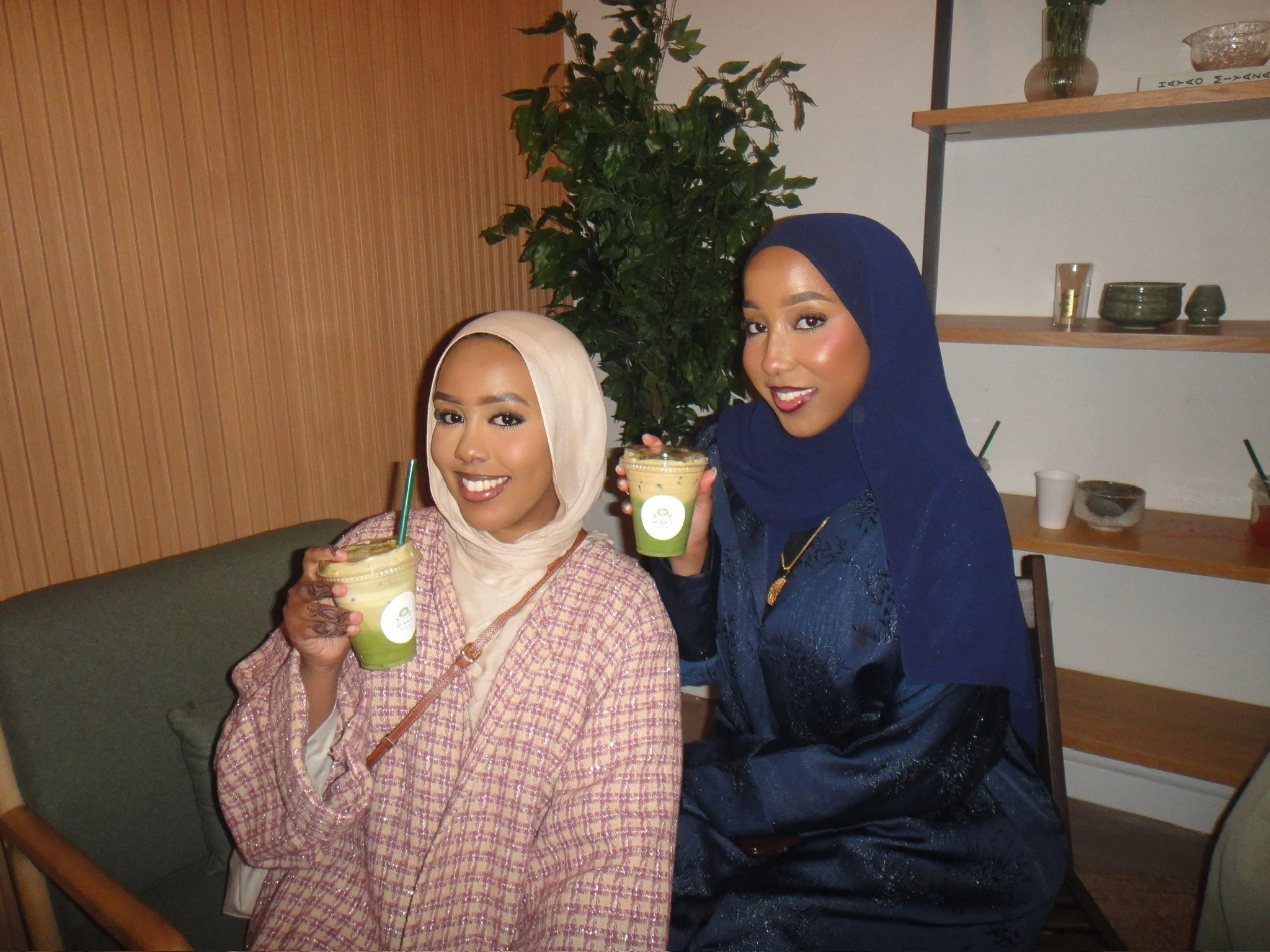 Two women of East African descent wearing hijabs and fashionable clothing, smiling and holding cold beverages with straws, sitting in a cafe or restaurant setting.