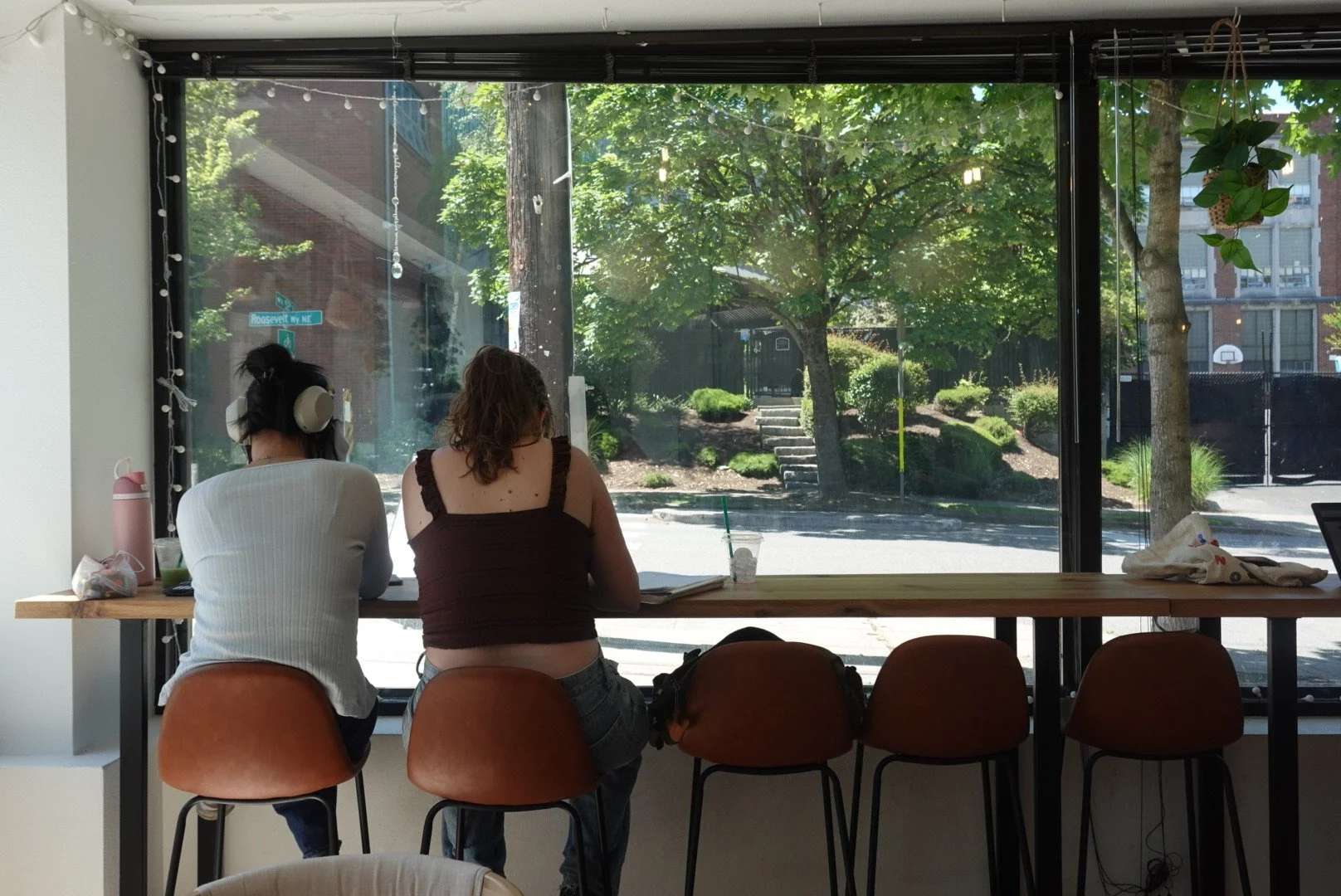 Two women are sitting at a wooden counter by a large window with a view of trees and a street. One woman has dark hair with headphones, and the other has curly hair. There are chairs around the counter, and personal items like a water bottle, a bag, 