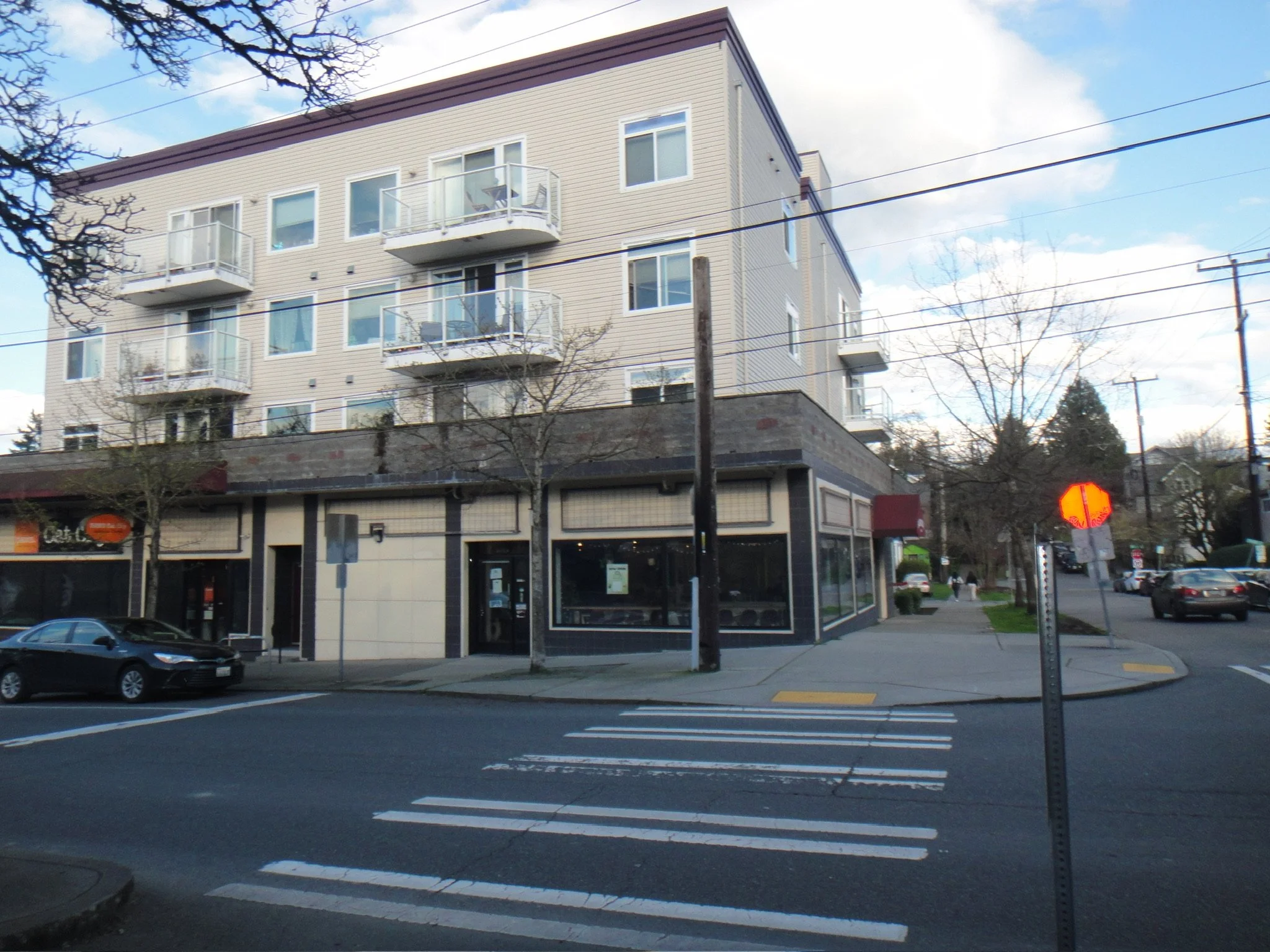 Corner of a three-story building with retail storefronts on the ground floor, beige siding on upper floors, and balconies with glass railings; parking lot with a car in front, and leafless trees along the sidewalk.