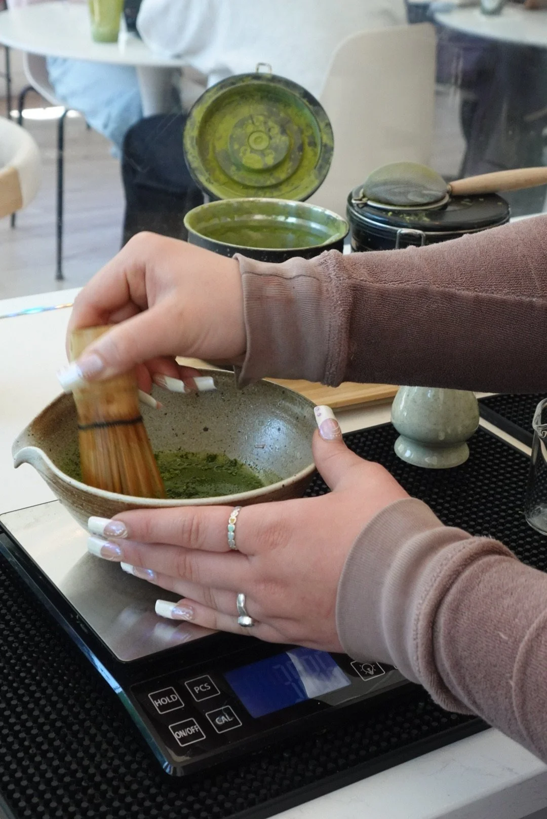 Person using a bamboo whisk to prepare matcha tea in a ceramic bowl, with several other matcha bowls and utensils on a table in the background.