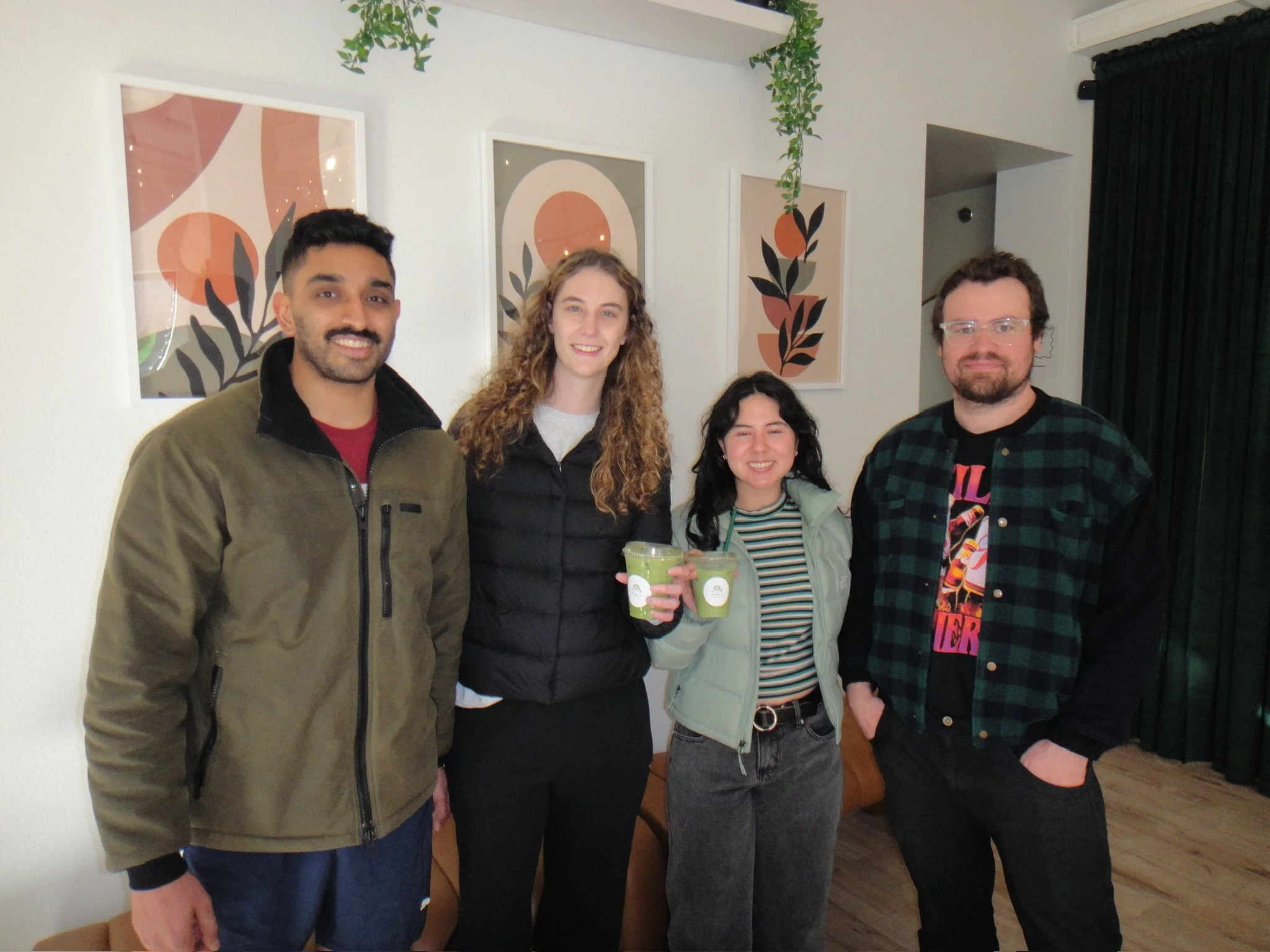 Group of five young adults smiling, standing indoors in a room with white walls and framed abstract art, two of them holding green smoothies.