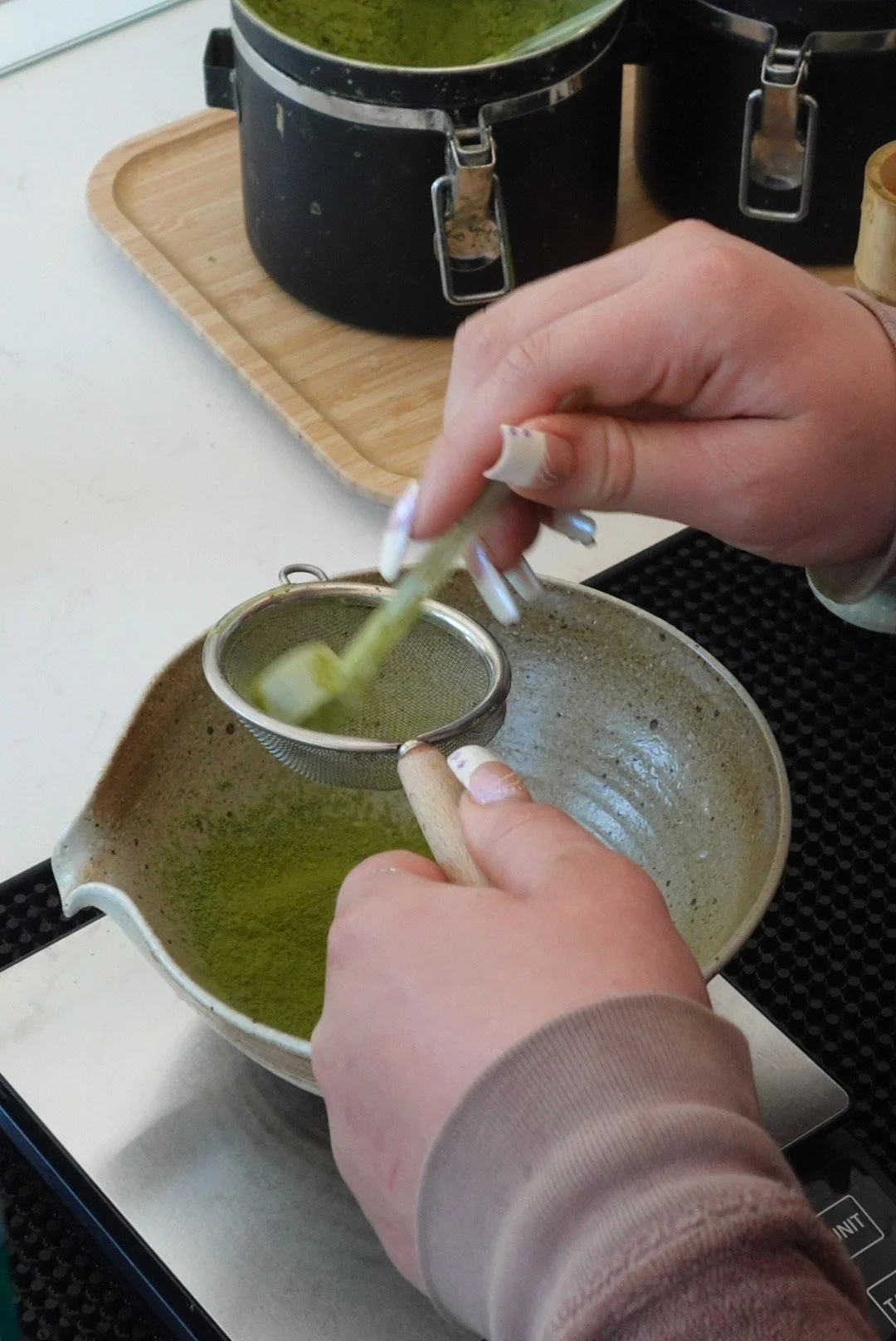Person straining green matcha tea with a fine mesh strainer over a ceramic bowl.