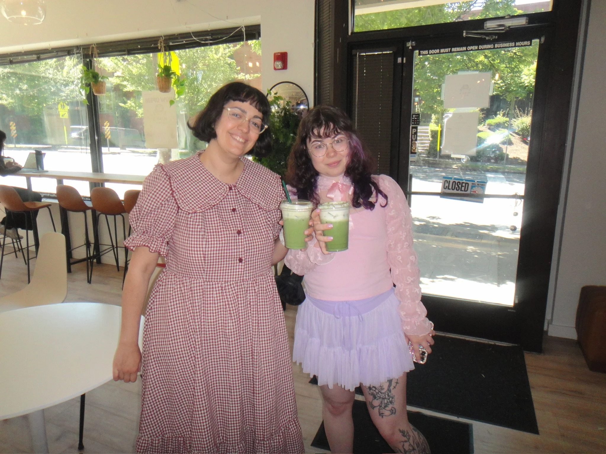 Two women standing together inside a cafe, holding green smoothies and smiling. One wearing a red and white checkered dress, the other wearing a pink lace top and a skirt.
