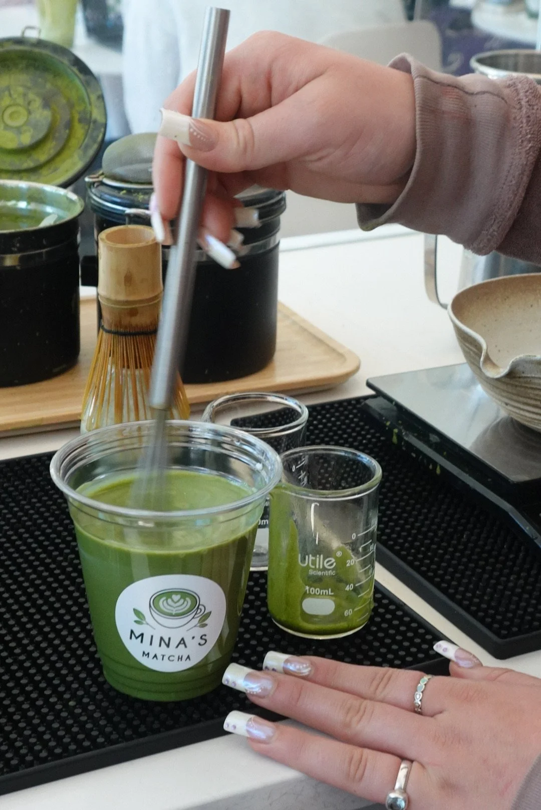 Close-up of a person's hand whisking bright green matcha tea into a clear plastic cup with a logo that reads "Mina's Matcha." The person has long, decorated nails and several rings. There are small glass beakers with matcha, a bamboo whisk, and other containers with matcha in the background on a black mat.