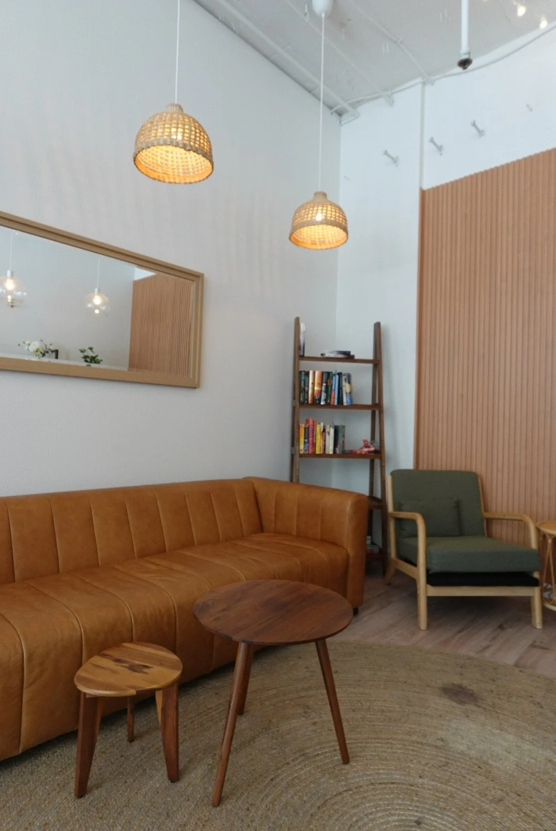 Cozy living room with a brown leather sofa, green armchair, wooden coffee table, bookshelf, large mirror, hanging wicker lamps, and a circular woven rug.