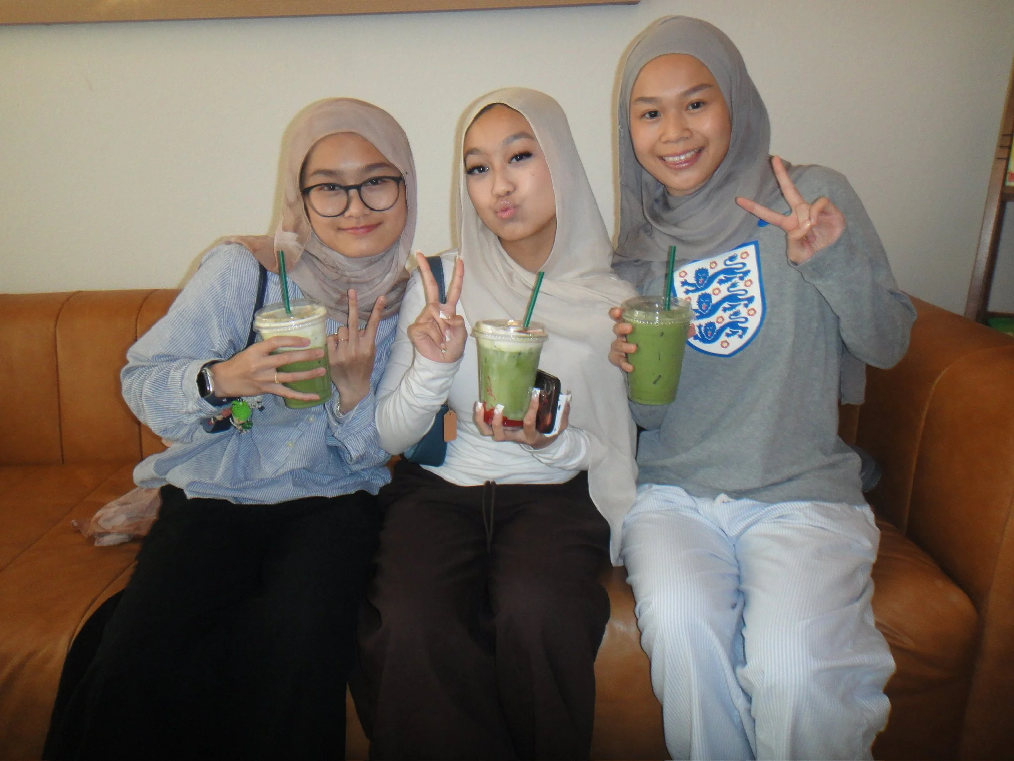 Three young women sitting on a brown leather sofa, smiling and posing with peace signs, each holding a green beverage in clear plastic cups with green straws, against a plain white wall background.