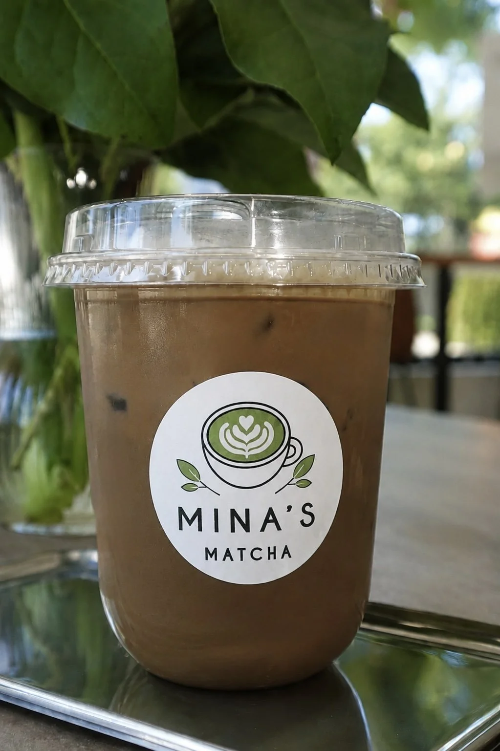 A plastic cup of iced hojicha latte with a lid, placed on a tray, from Mina's Matcha, with a leafy plant and blurred outdoor background.