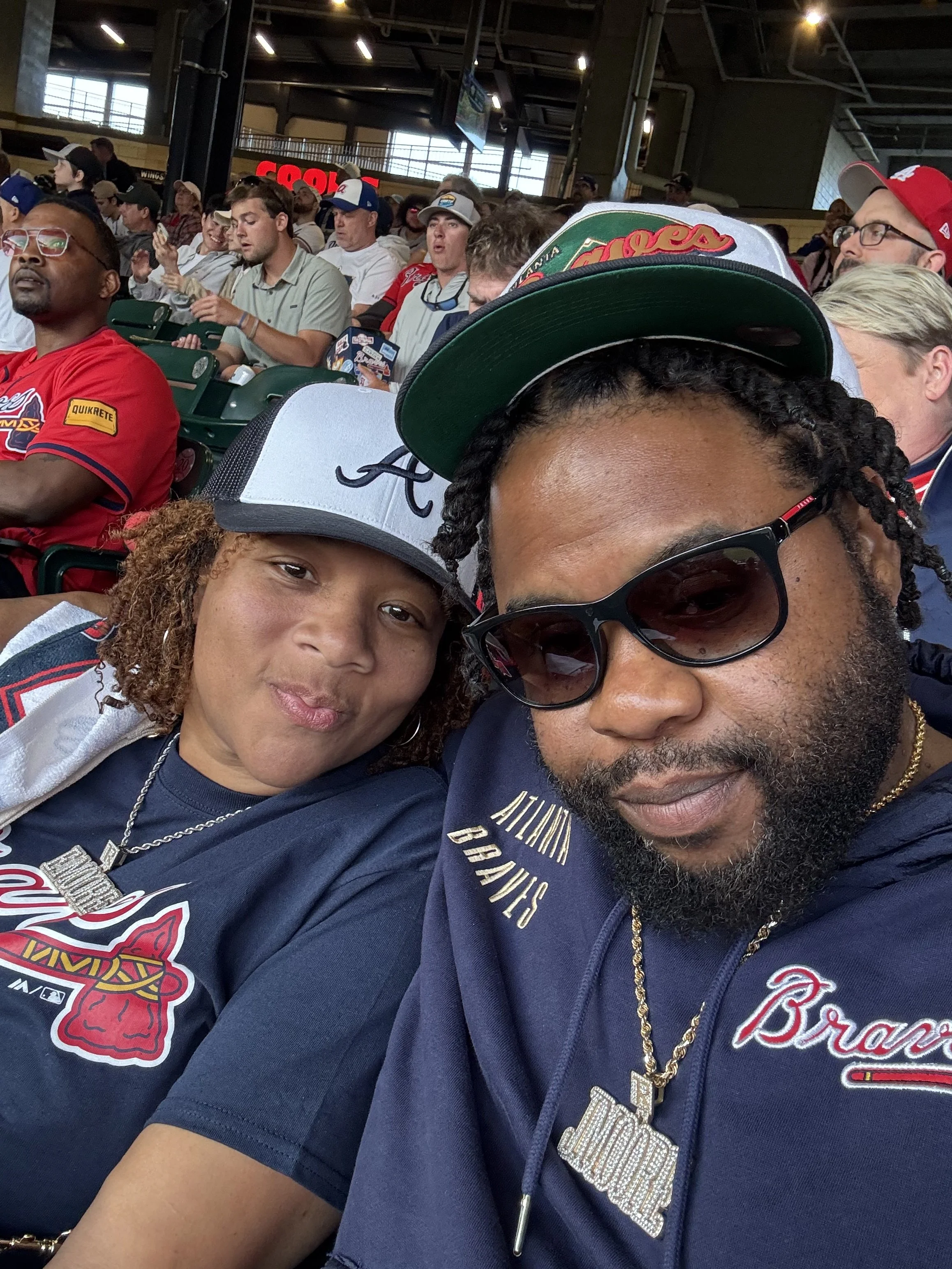 Two people taking a selfie at a sports stadium filled with fans, some wearing Atlanta Braves apparel, with a large crowd in the background.