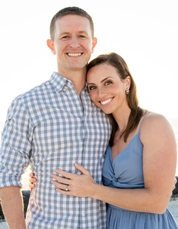 A smiling couple standing outdoors, the man wearing a checkered shirt and the woman in a blue dress, embracing each other with the ocean in the background.