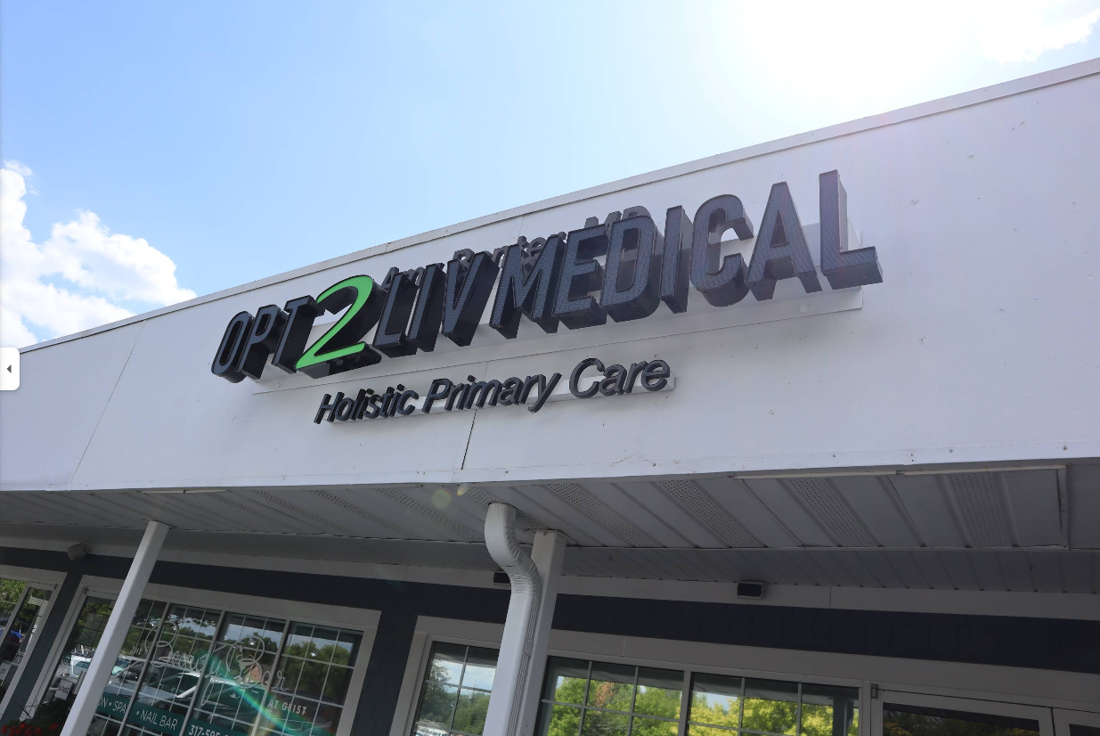 Exterior view of OPT2 LIV MEDICAL, a holistic primary care clinic, showing the sign on the building's white facade against a partly cloudy sky.