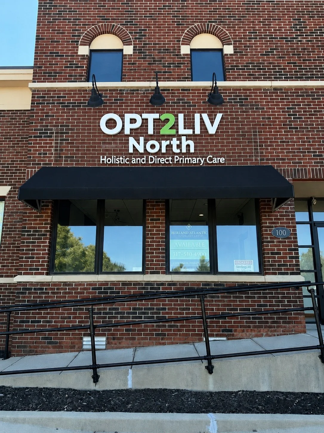 Exterior of a brick building with the sign 'OPT 2 LIV North' and the phrase 'Holistic and Direct Primary Care' underneath. A black awning covers the entrance, and windows display a sign for available medical services and a contact number.
