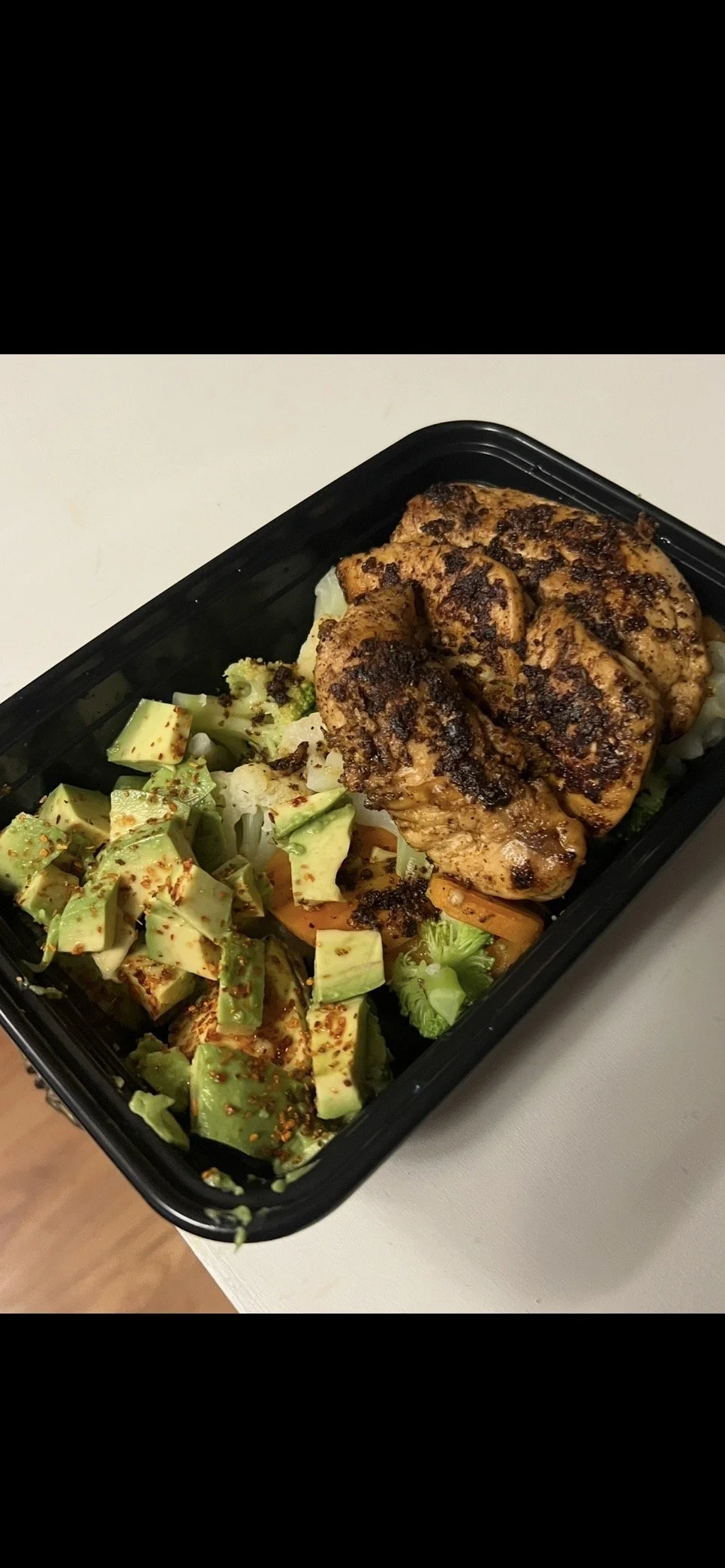 Grilled chicken breast with charred marks served with chopped avocado and vegetables in a black takeout container.