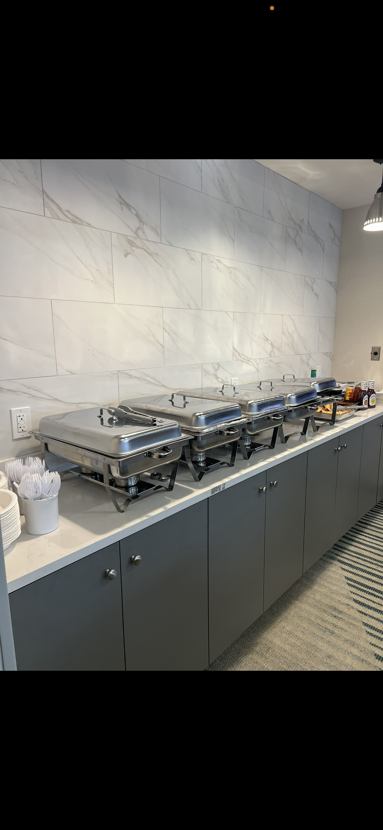 Buffet station with five chafing dishes on a marble countertop in a dining area, with condiment bottles, utensils, and disposable cups and plates nearby.