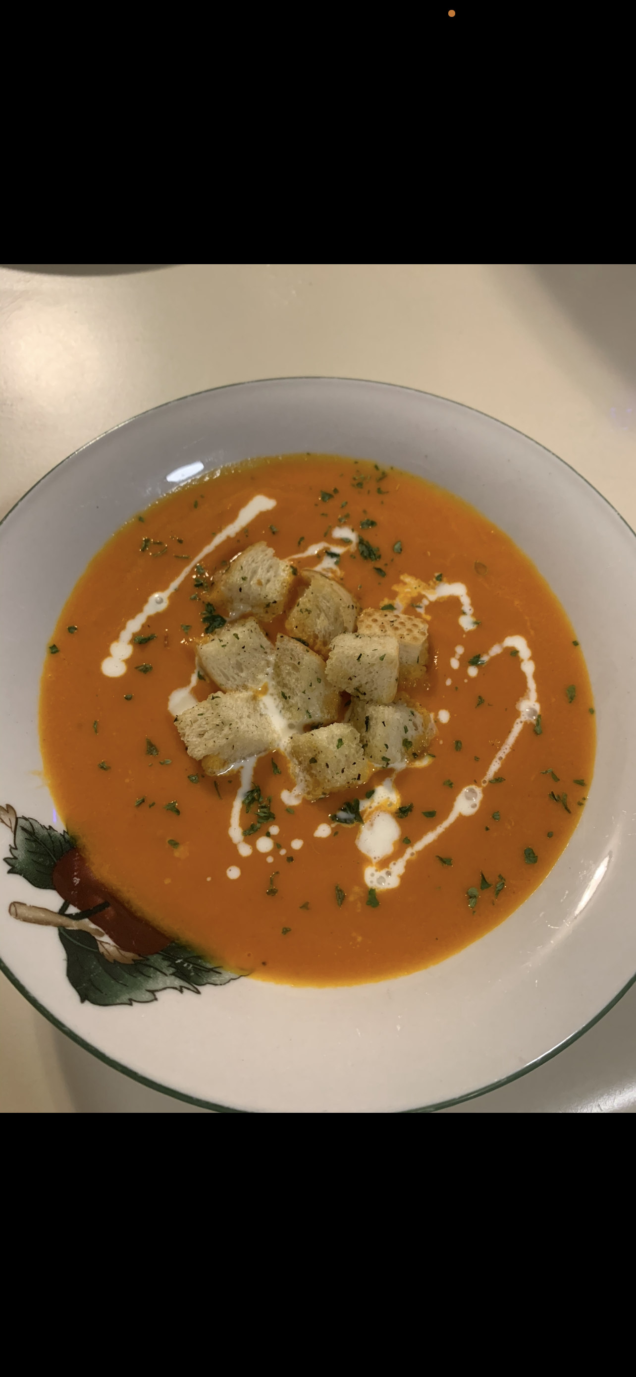 A bowl of orange soup garnished with croutons, a drizzle of white sauce, and chopped herbs.
