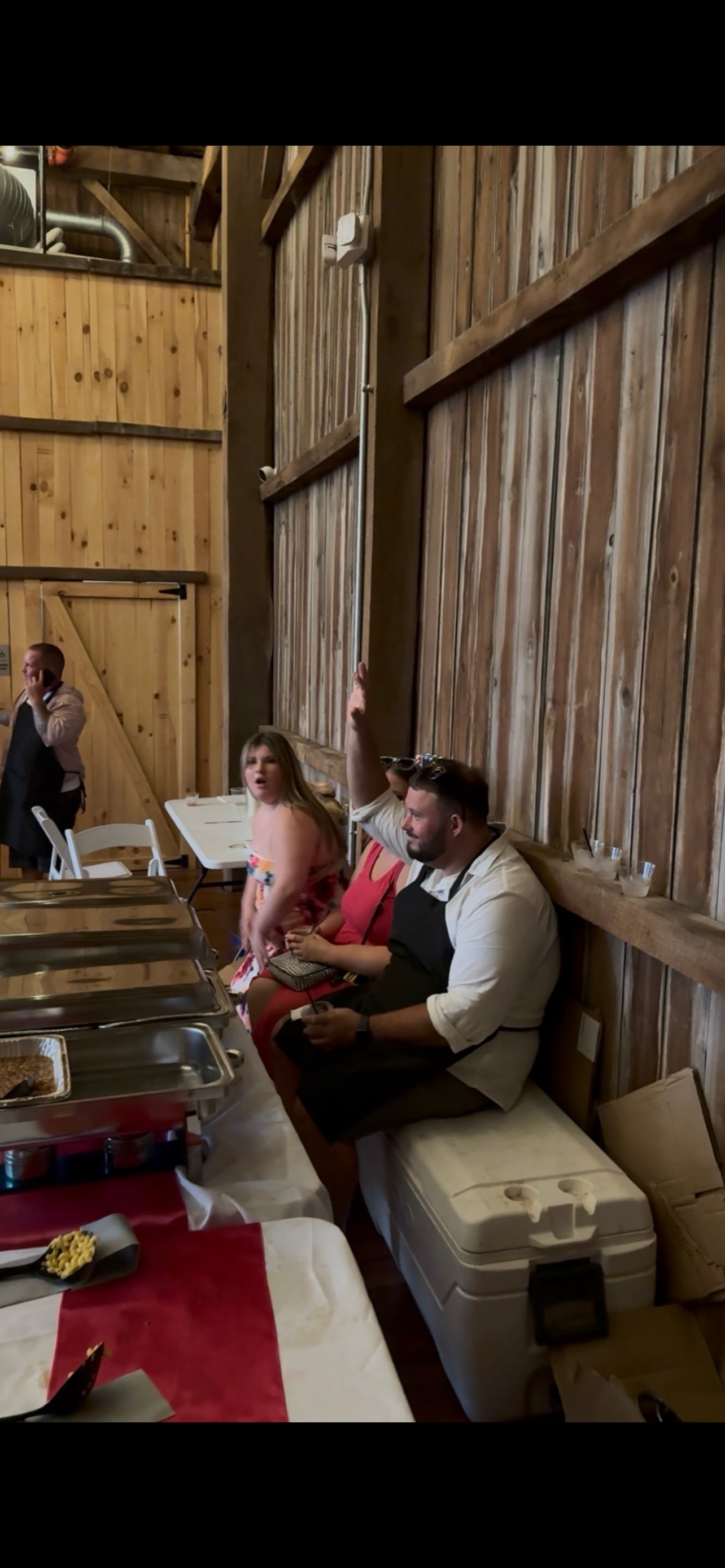 People sitting at a table in a wooden-finished room, with one woman raising her hand, a man sitting holding a smartphone, and a young girl standing next to them.