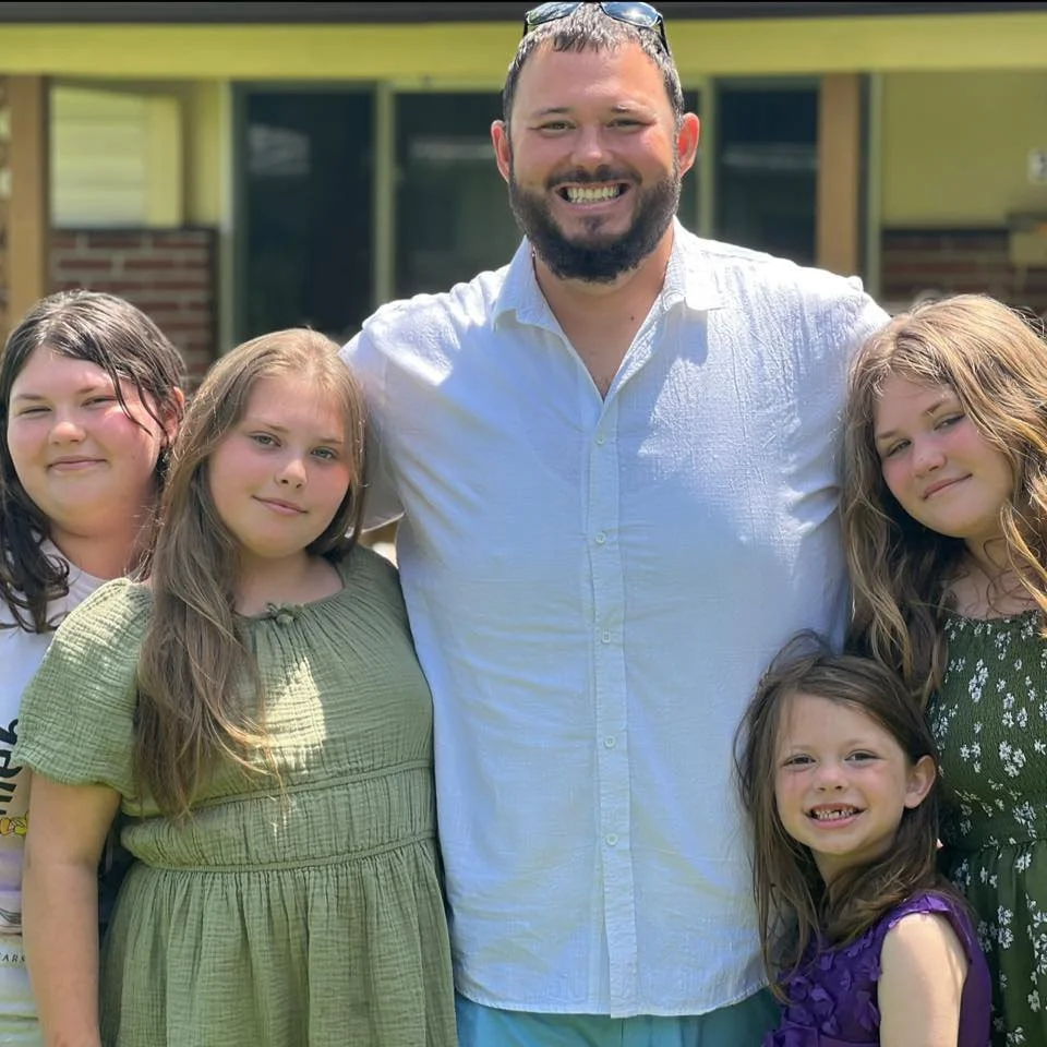 A man with a beard and glasses on his head standing outdoors with four young girls, all smiling and posing for the photo.