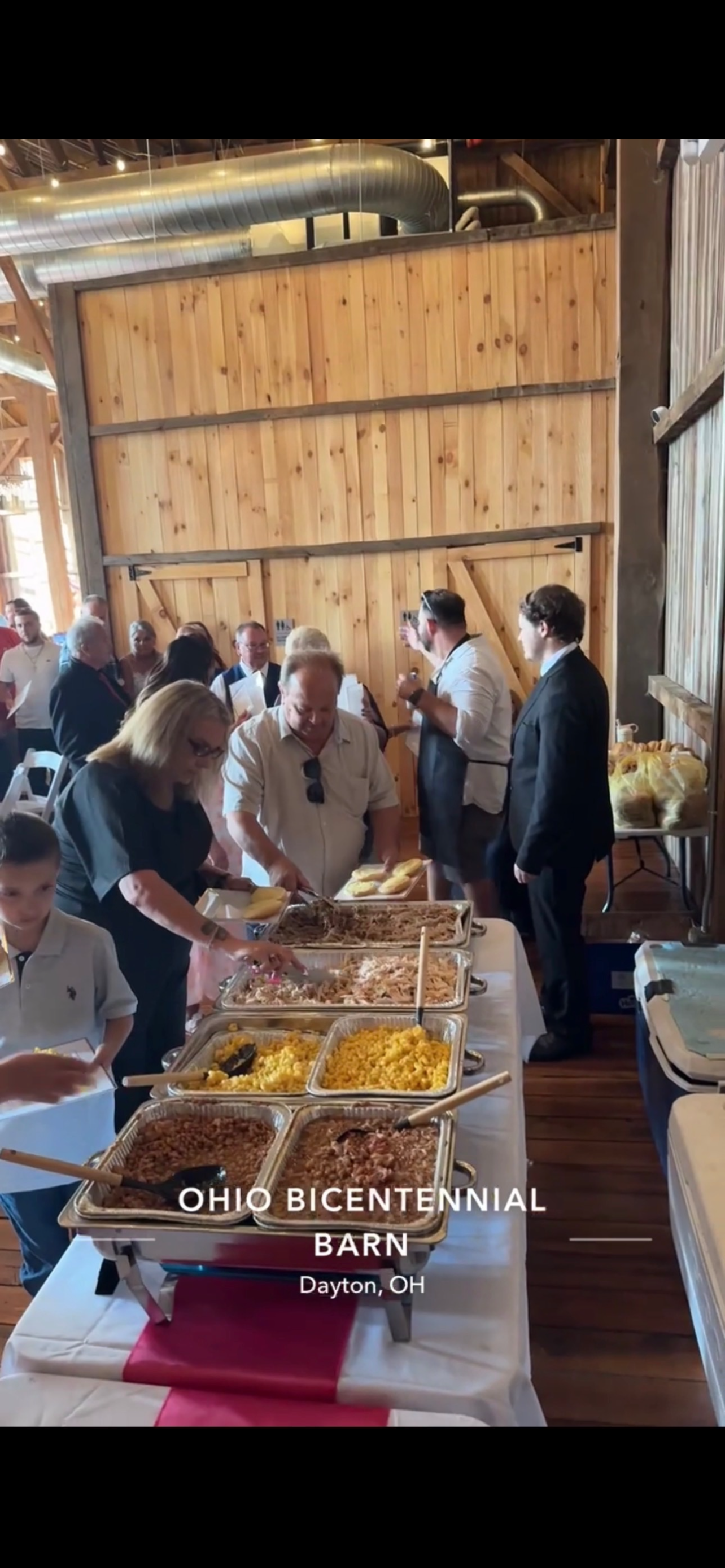 People gathering around a buffet table with various dishes at Ohio Bicentennial Barn event in Dayton, OH, inside a wooden barn with large air ducts and wooden walls.