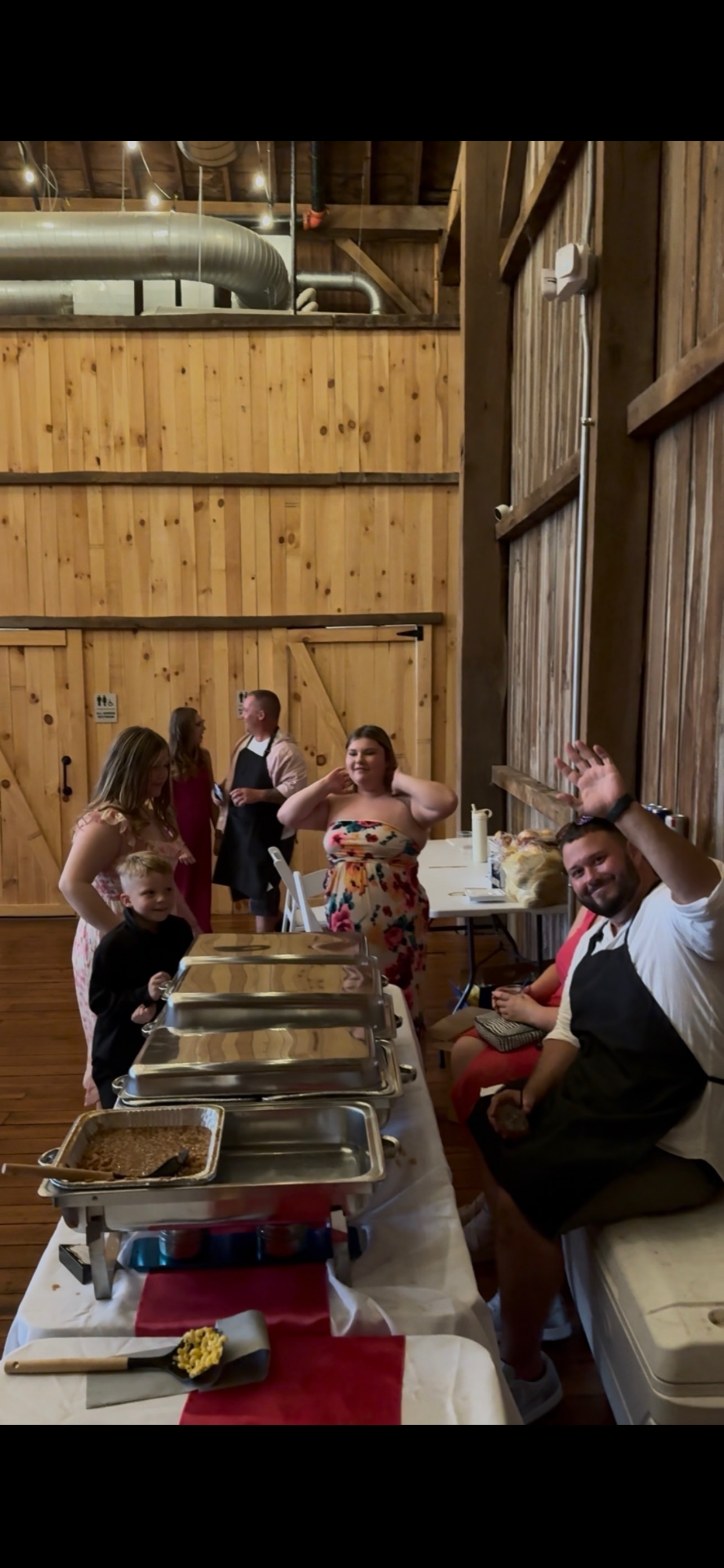People gathered in a rustic wooden room with buffet-style food serving station, some smiling and waving at the camera, celebrating an event or gathering.