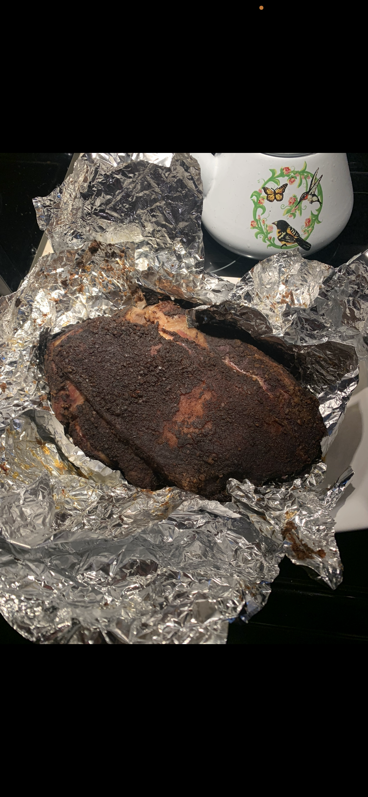 A cooked piece of meat, likely a brisket, with a dark bark crust, wrapped in crumpled aluminum foil.