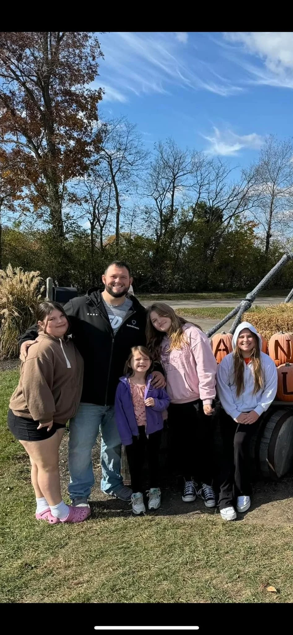 A group of five people, including three young girls, a man, and a woman, posing outdoors during daytime with a blue sky, leafless trees, and Halloween decorations with pumpkins in the background.