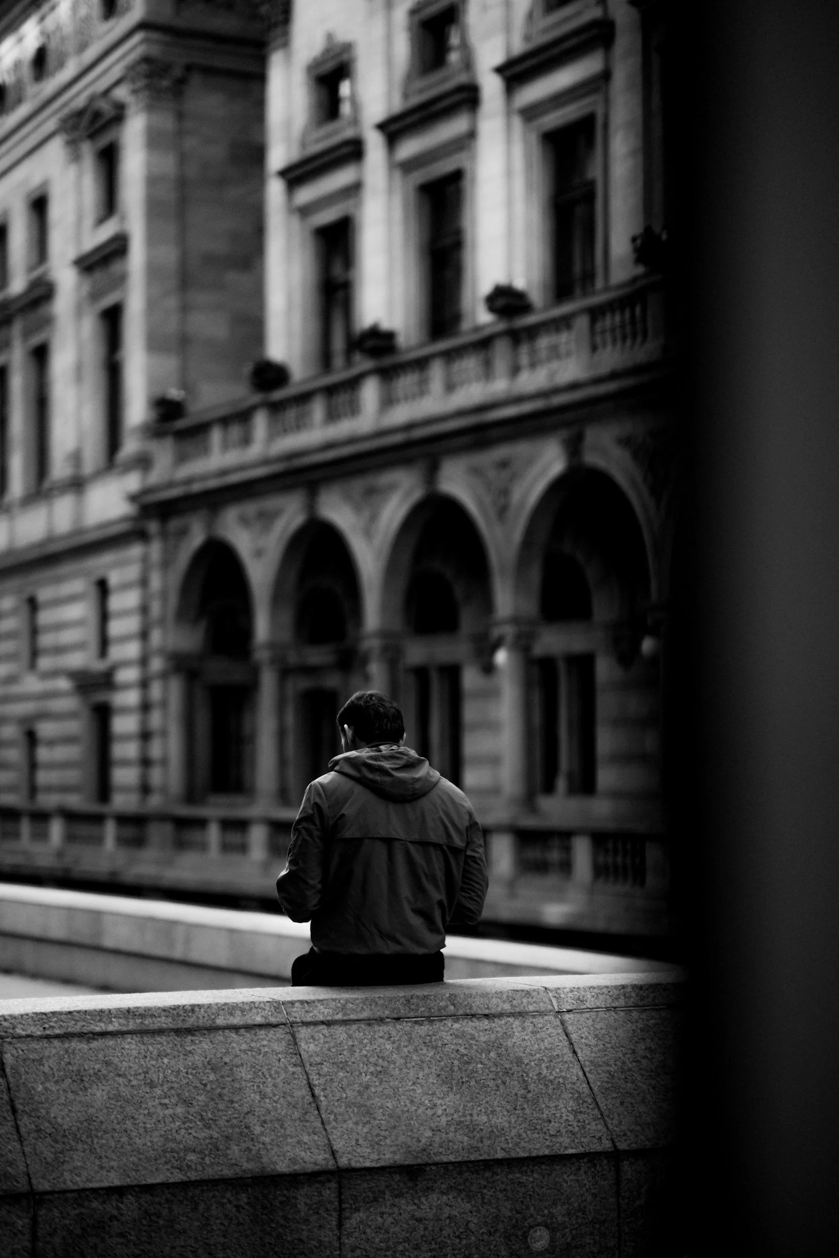 A person with short dark hair, wearing a hooded jacket, sitting alone on a curved stone ledge in a city area. In the background, there is a large, ornate building with multiple stories, arched windows, and decorative architectural elements, captured in black and white.