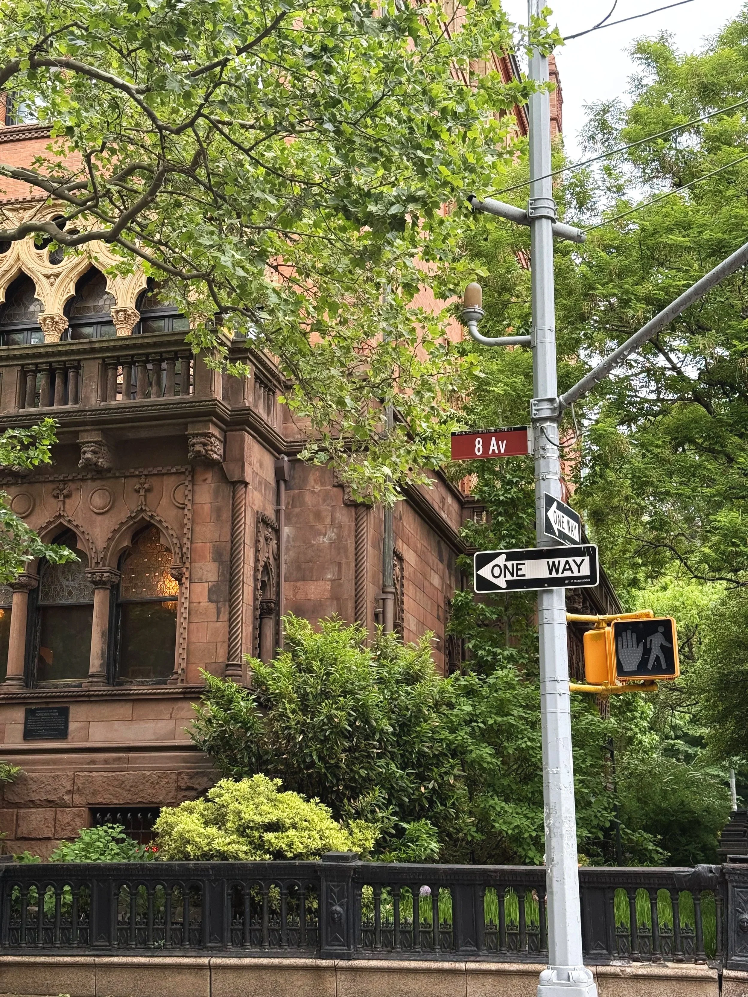 Street corner with decorative brownstone building, street signs indicating 8th Avenue, and pedestrian crossing signal, surrounded by green trees and bushes.