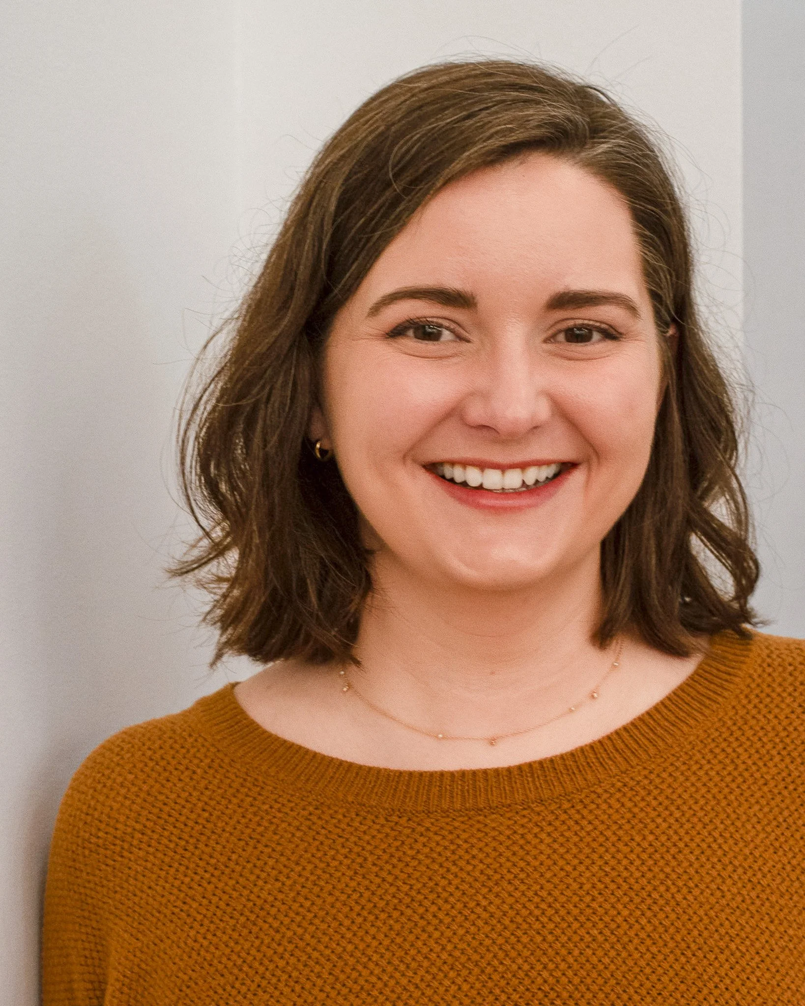 Close-up of a smiling woman with brown hair wearing a mustard sweater and a delicate gold necklace, standing against a plain light-colored background.