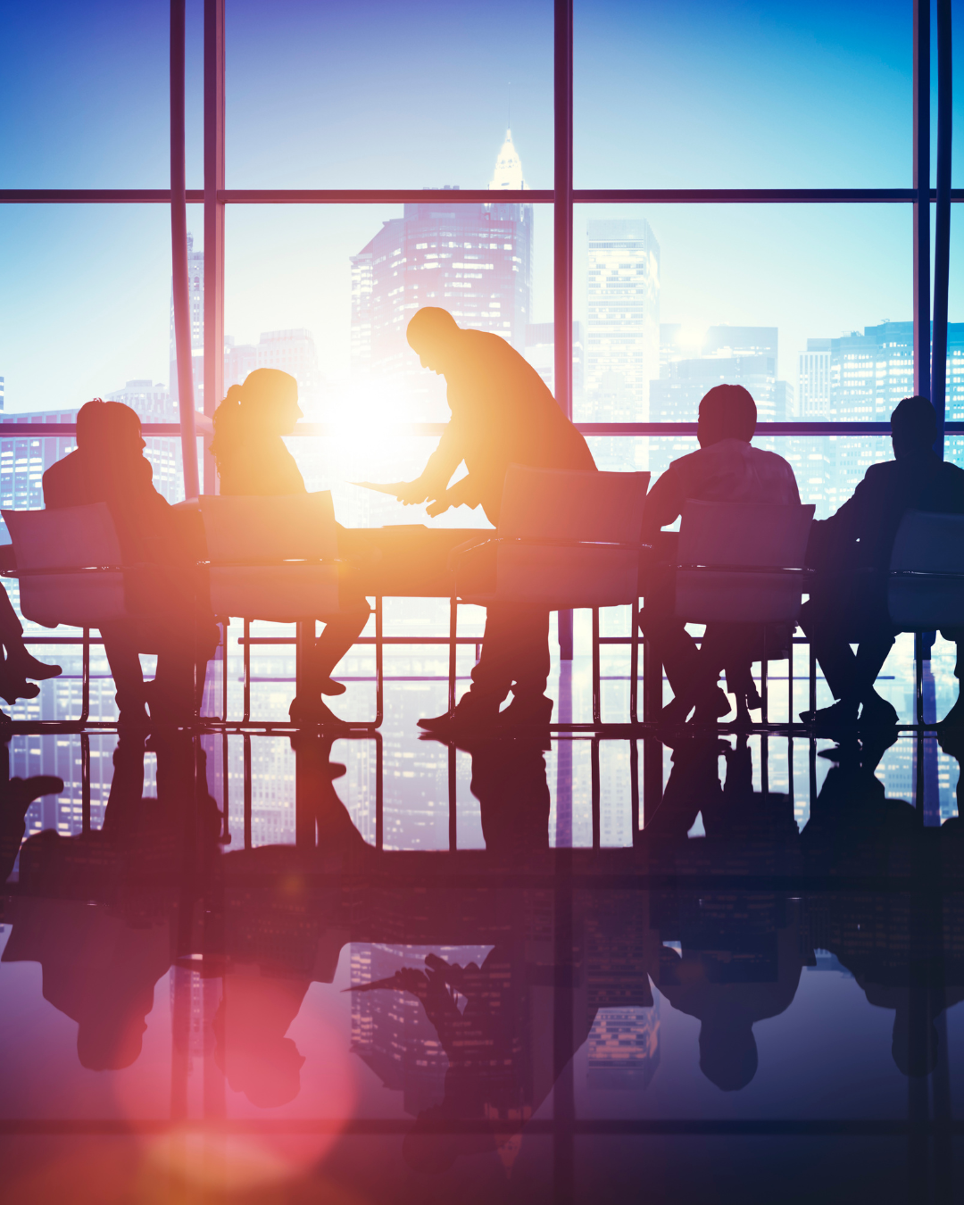 Silhouettes of six people in a business meeting or conference room with large windows showing a city skyline and tall buildings in the background, during sunrise or sunset.