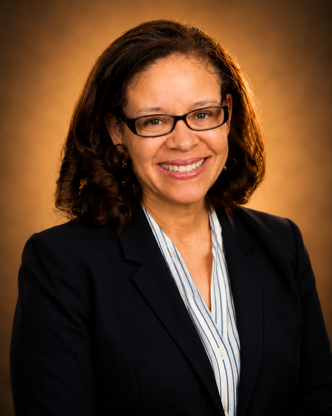 Professional woman with glasses, curly hair, wearing a dark blazer and striped blouse, smiling against a brown background.
